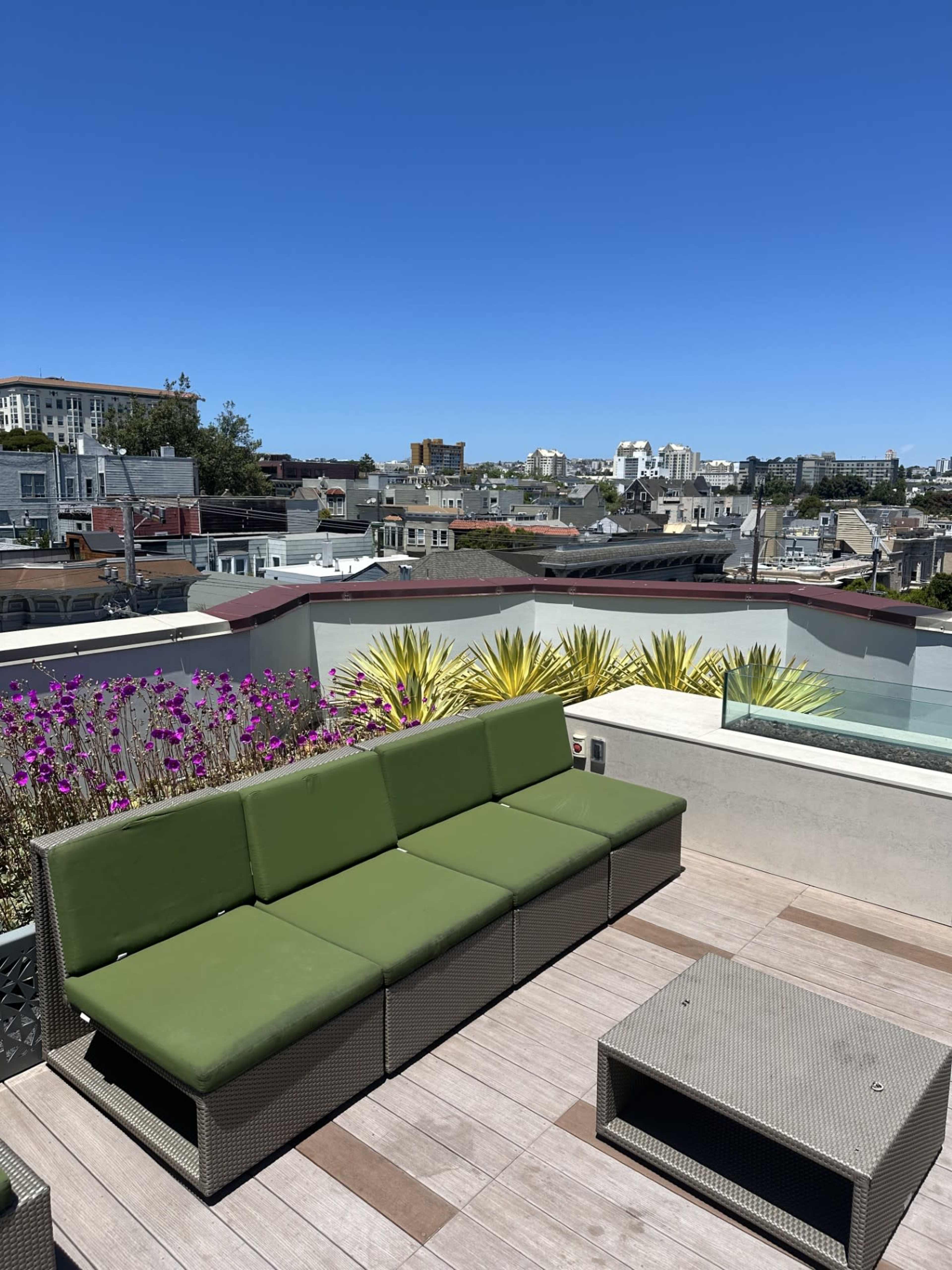A rooftop lounge area features a green sectional sofa and a small coffee table, overlooking a city skyline under a clear blue sky.