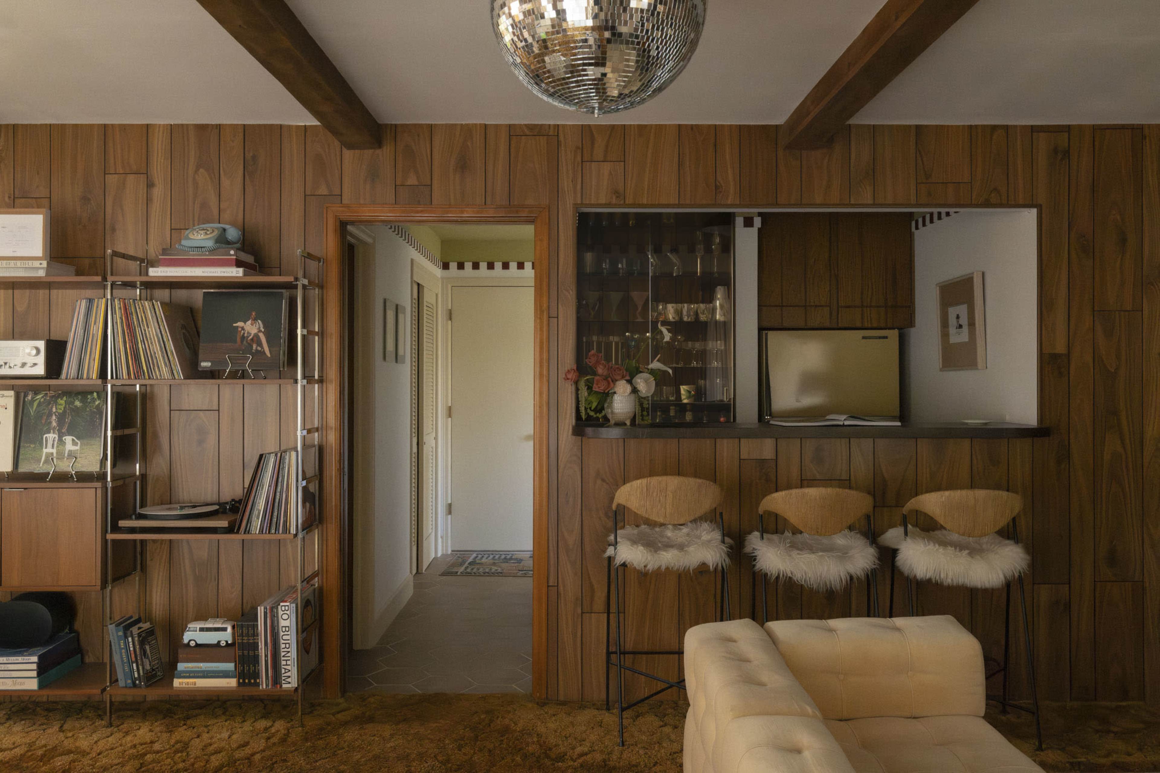 The image shows a mid-century modern interior with wooden paneling, a bar area featuring three stools, and a collection of records displayed on shelves.