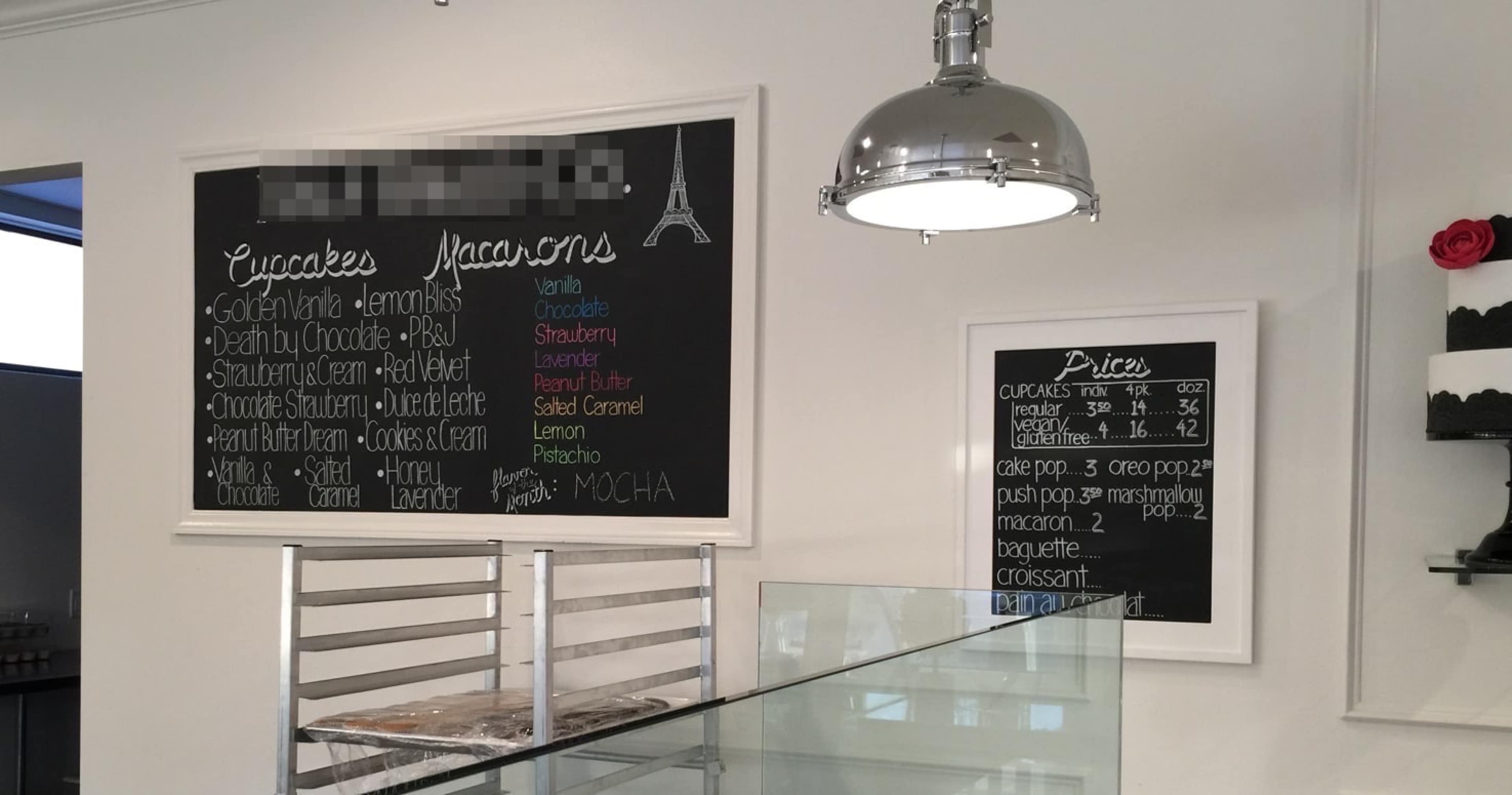 A menu board displays various flavors of cupcakes and macarons in a bakery setting.