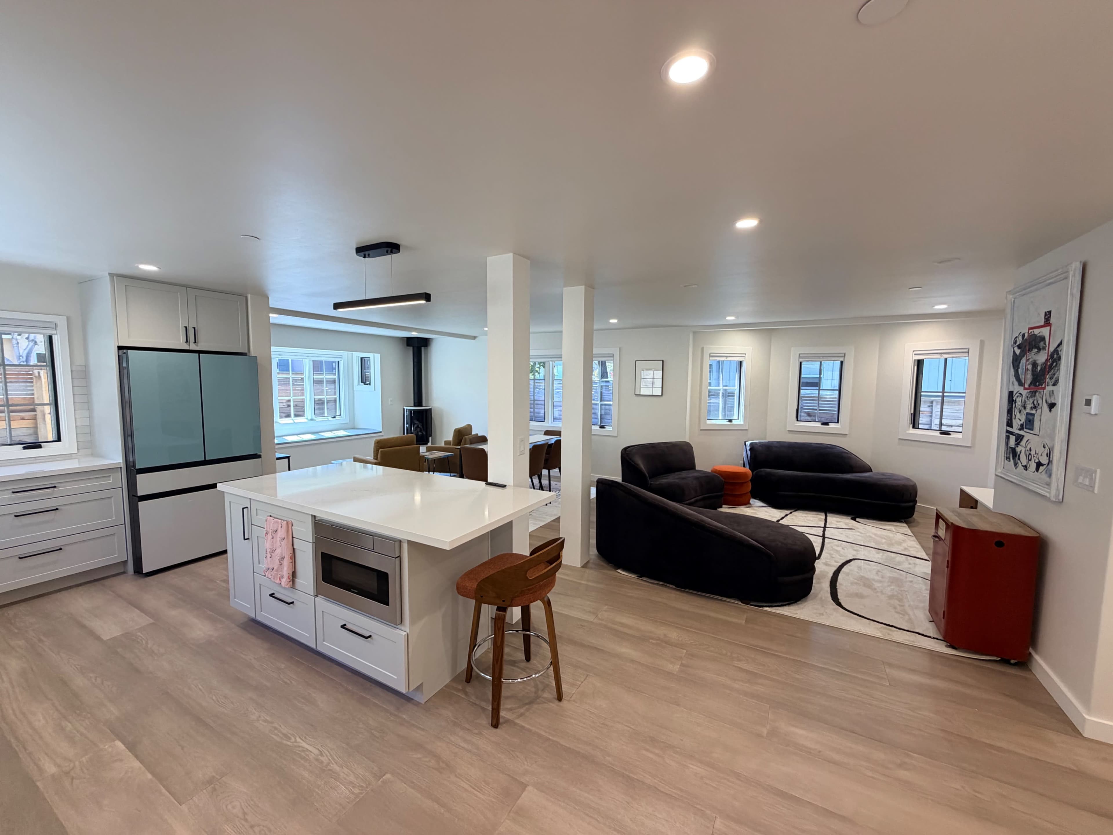 The image shows a modern, open-concept living area with a kitchen island, a seating area featuring dark chairs and a coffee table, and large windows letting in natural light.