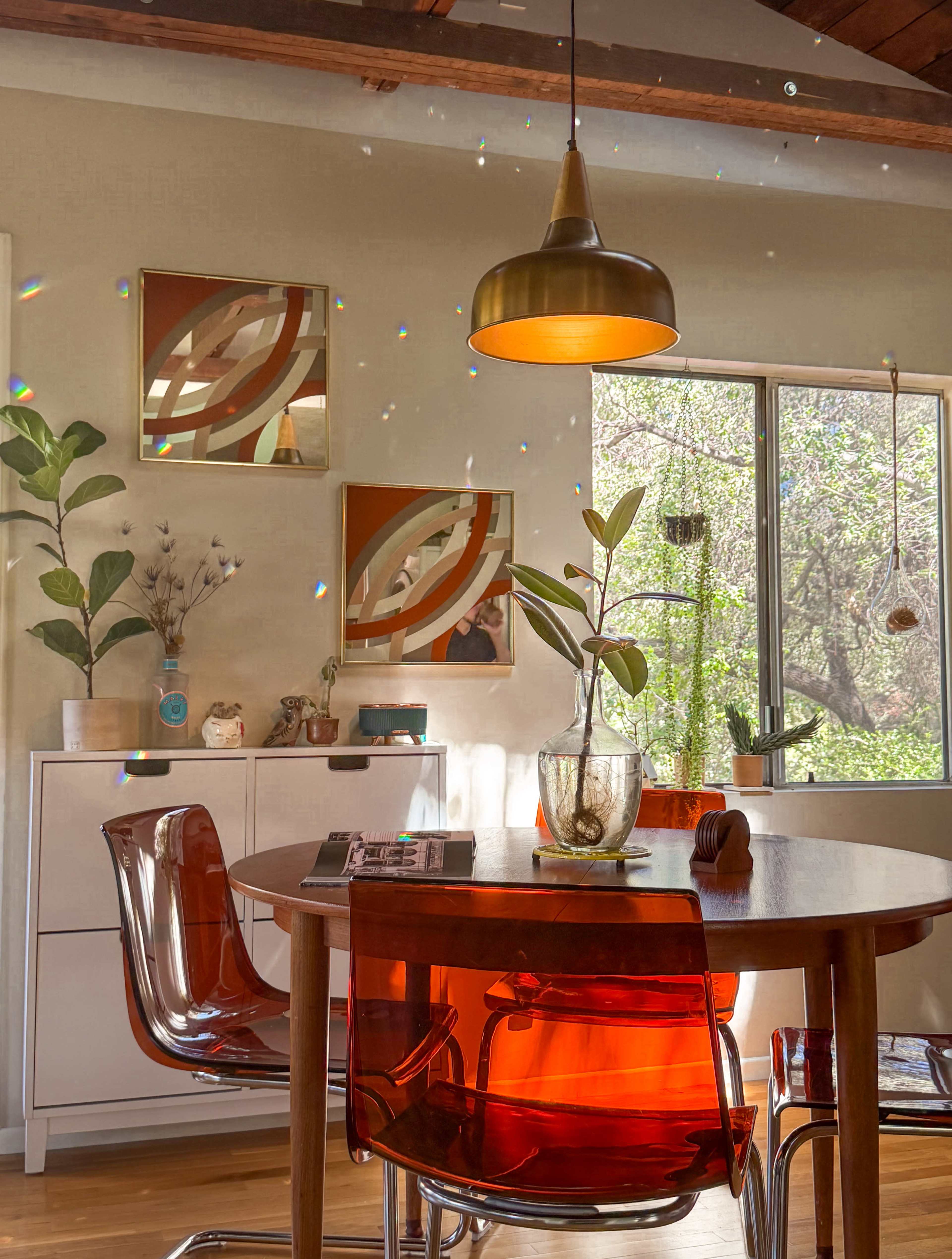 A dining area features a round wooden table surrounded by transparent orange chairs, with a large window providing natural light and displaying greenery outside.