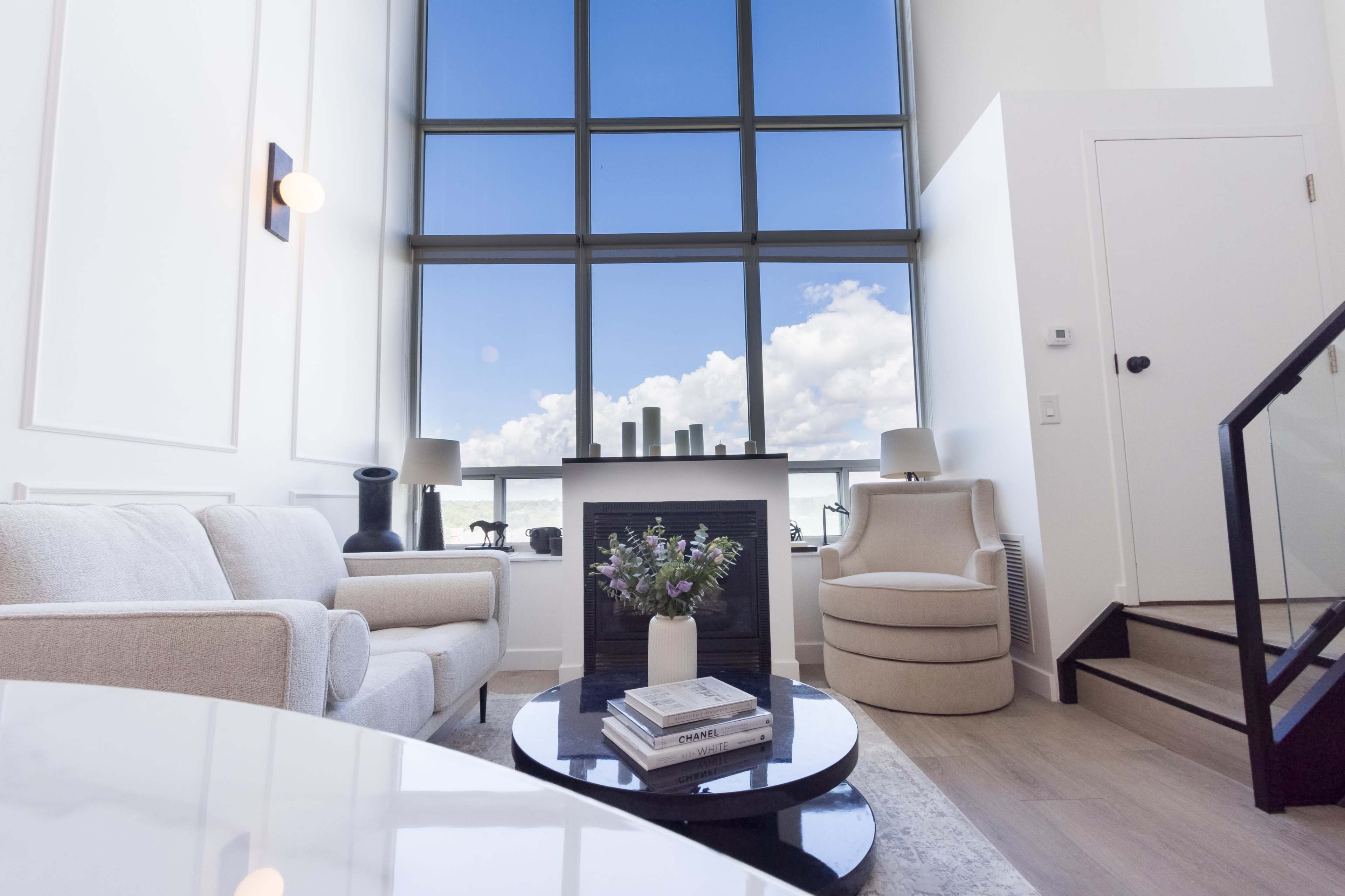 A modern living room features a large window with views of blue skies and clouds, a white sofa, a round chair, and a coffee table with books and a vase of flowers.