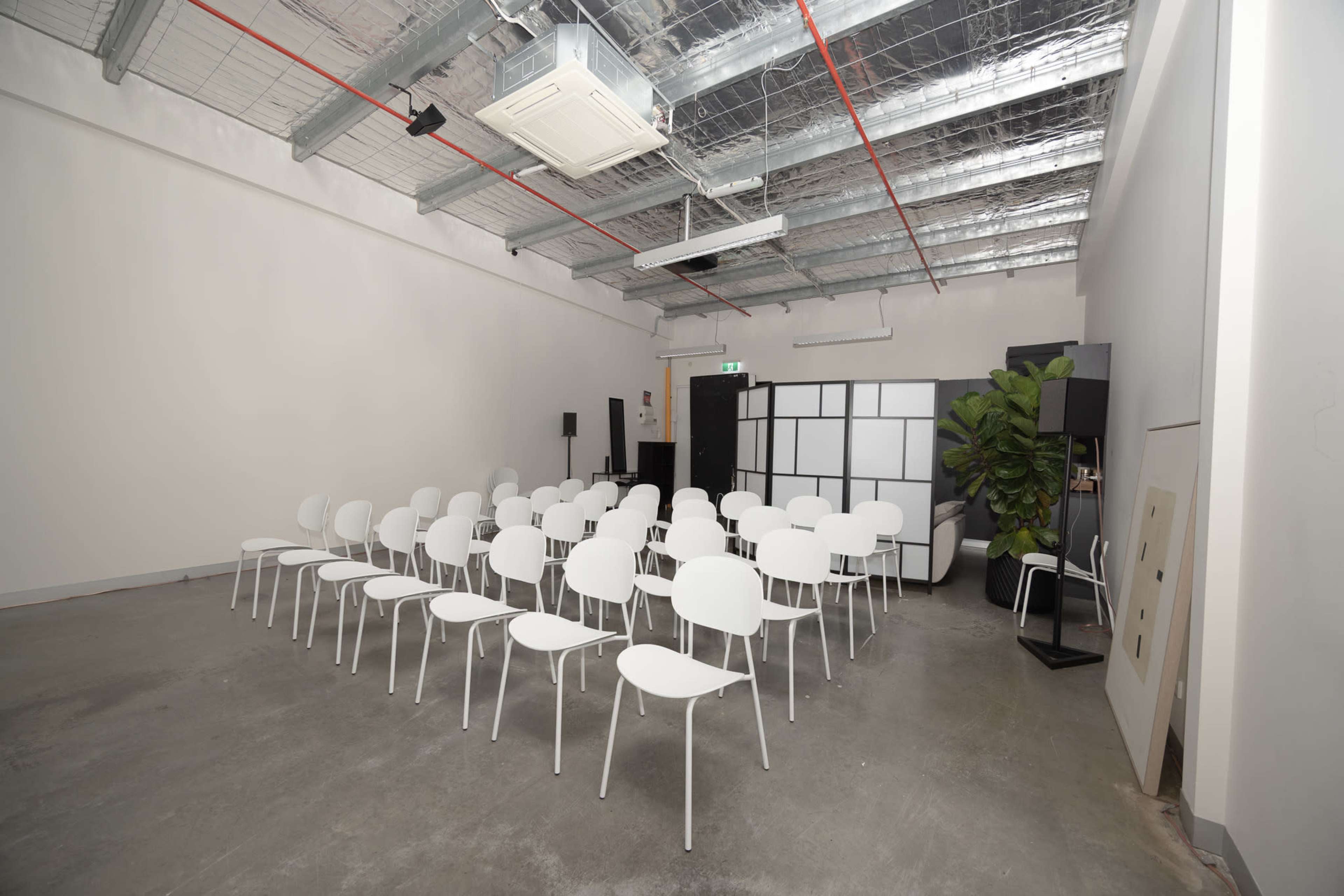 The image shows a minimalist meeting room with rows of white chairs facing a partition and a potted plant in the corner.