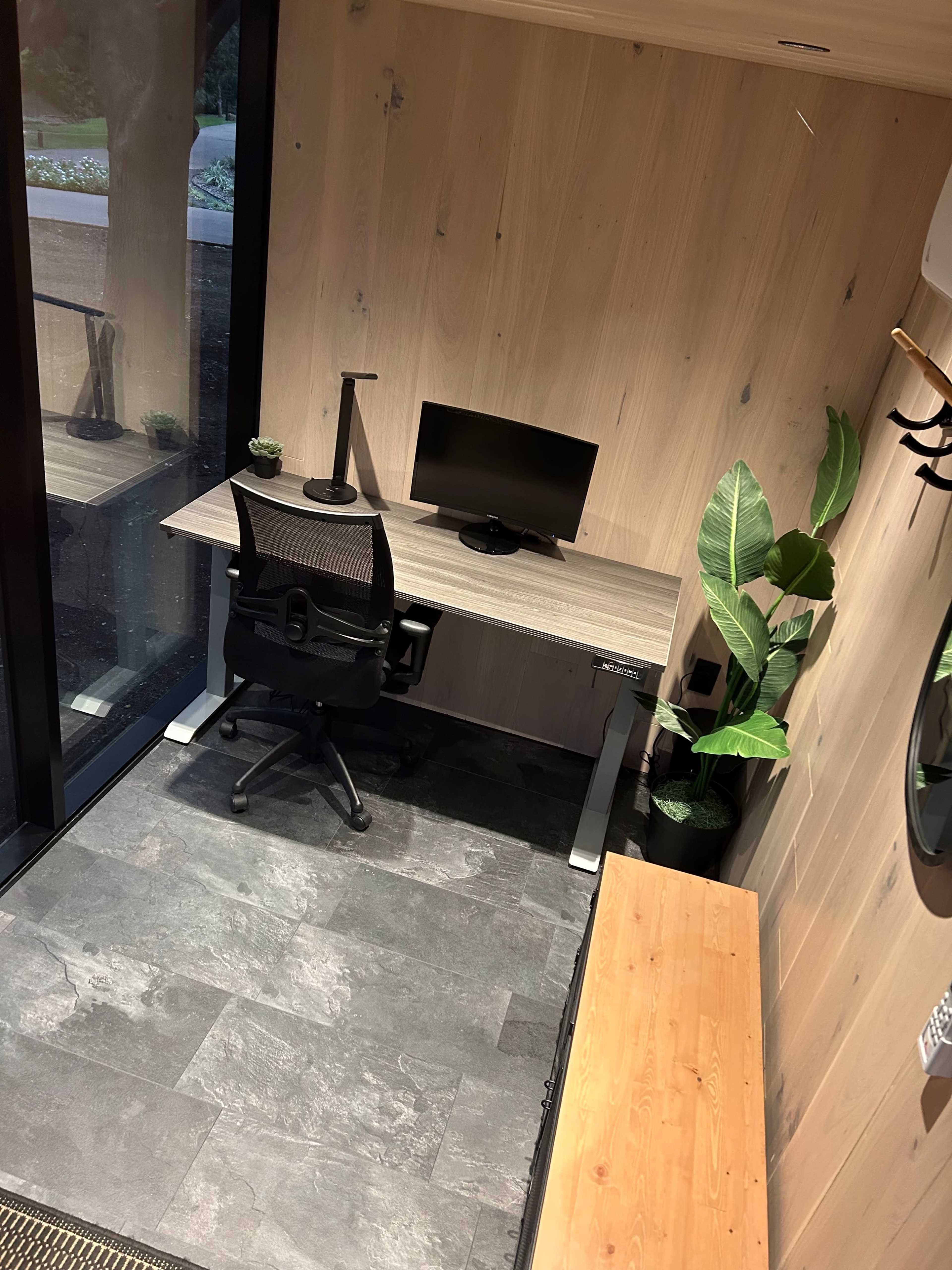 The image shows a minimalist workspace featuring a desk with a computer monitor, an office chair, and decorative plants in a room with wooden walls and stone flooring.