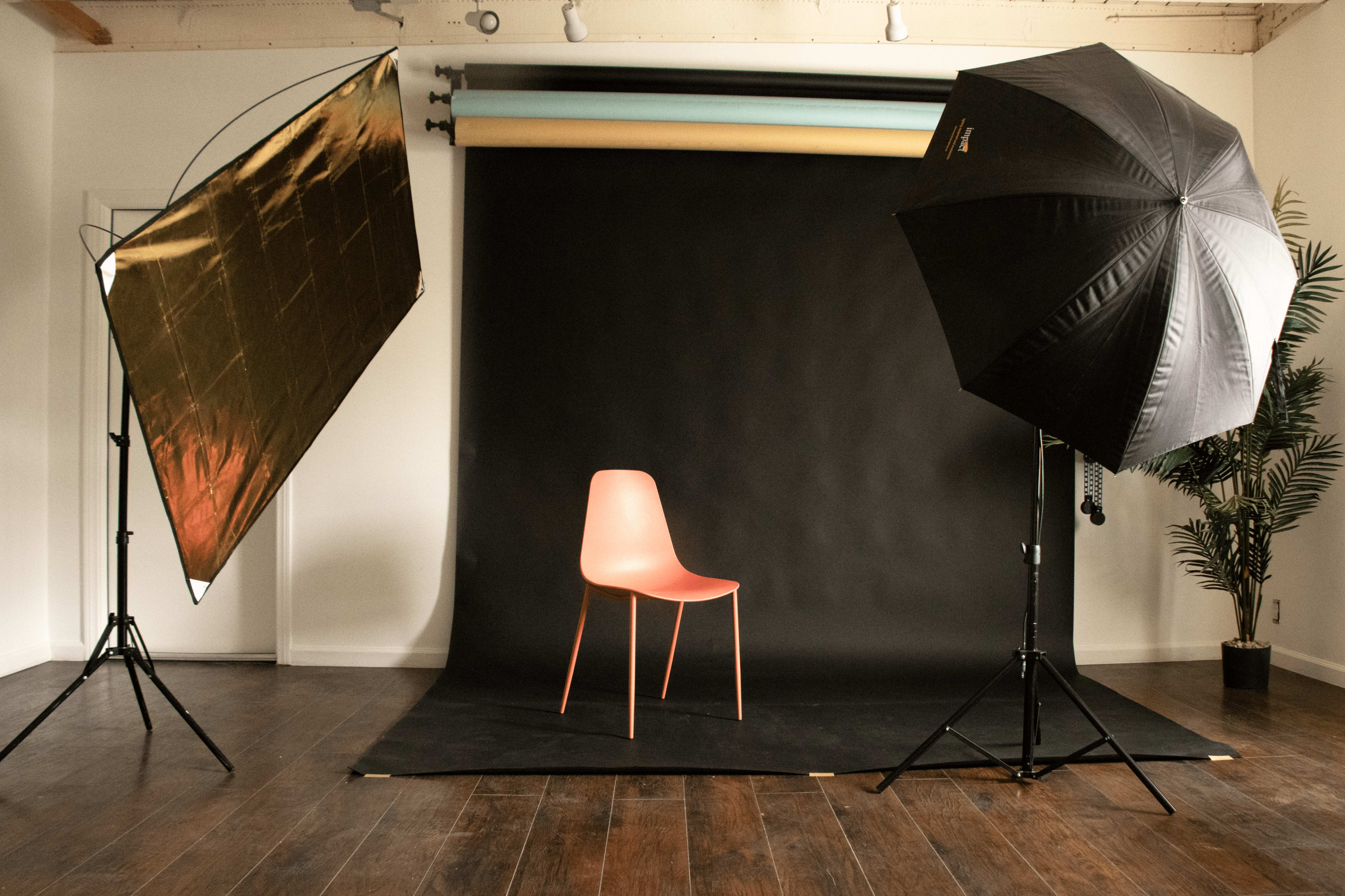 A pink chair is positioned on a dark backdrop in a photography studio, flanked by a gold reflector and a large black umbrella light.