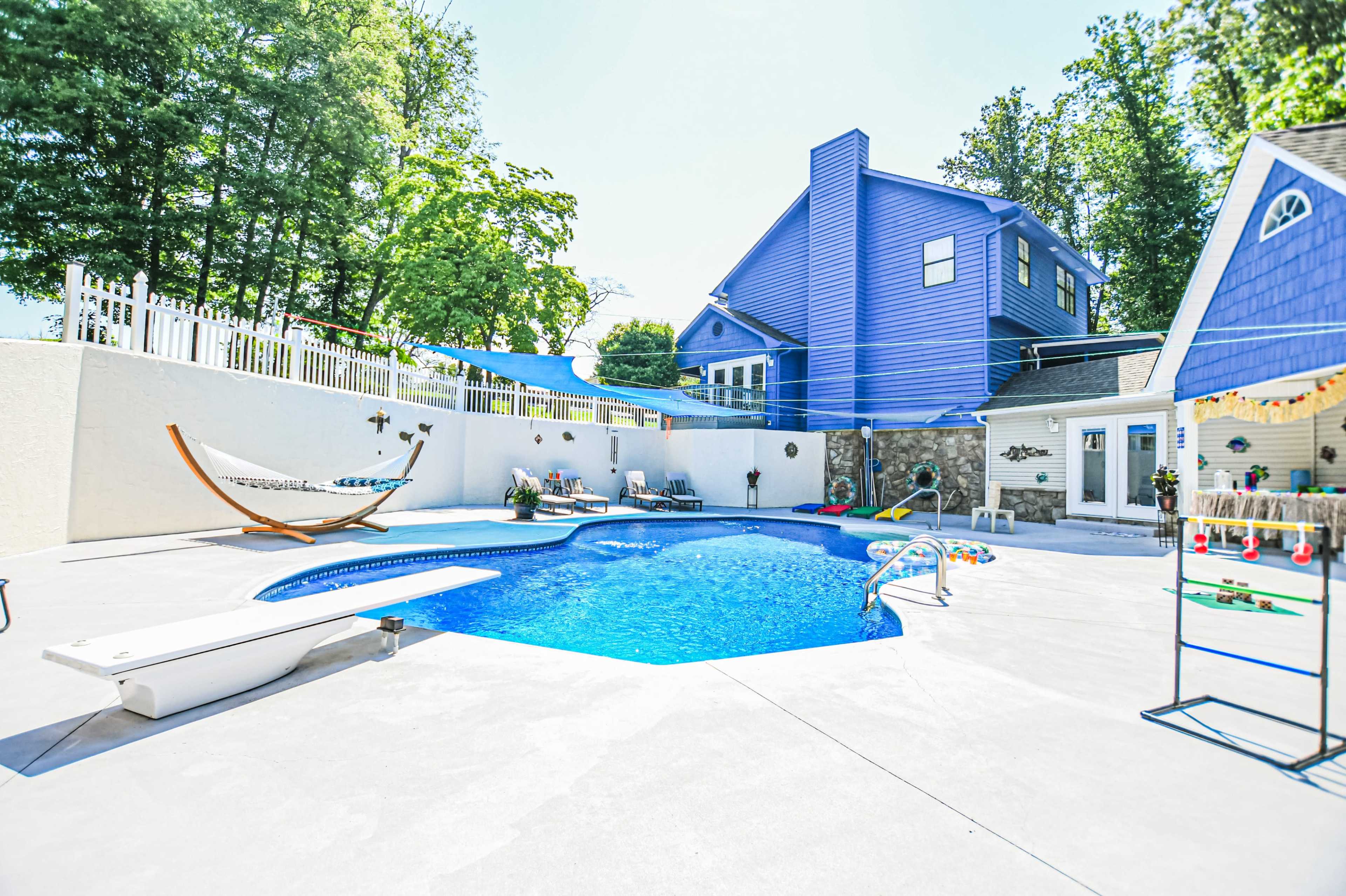 The image shows a backyard with a blue swimming pool surrounded by a concrete patio, a hammock, and a house with blue siding in the background.