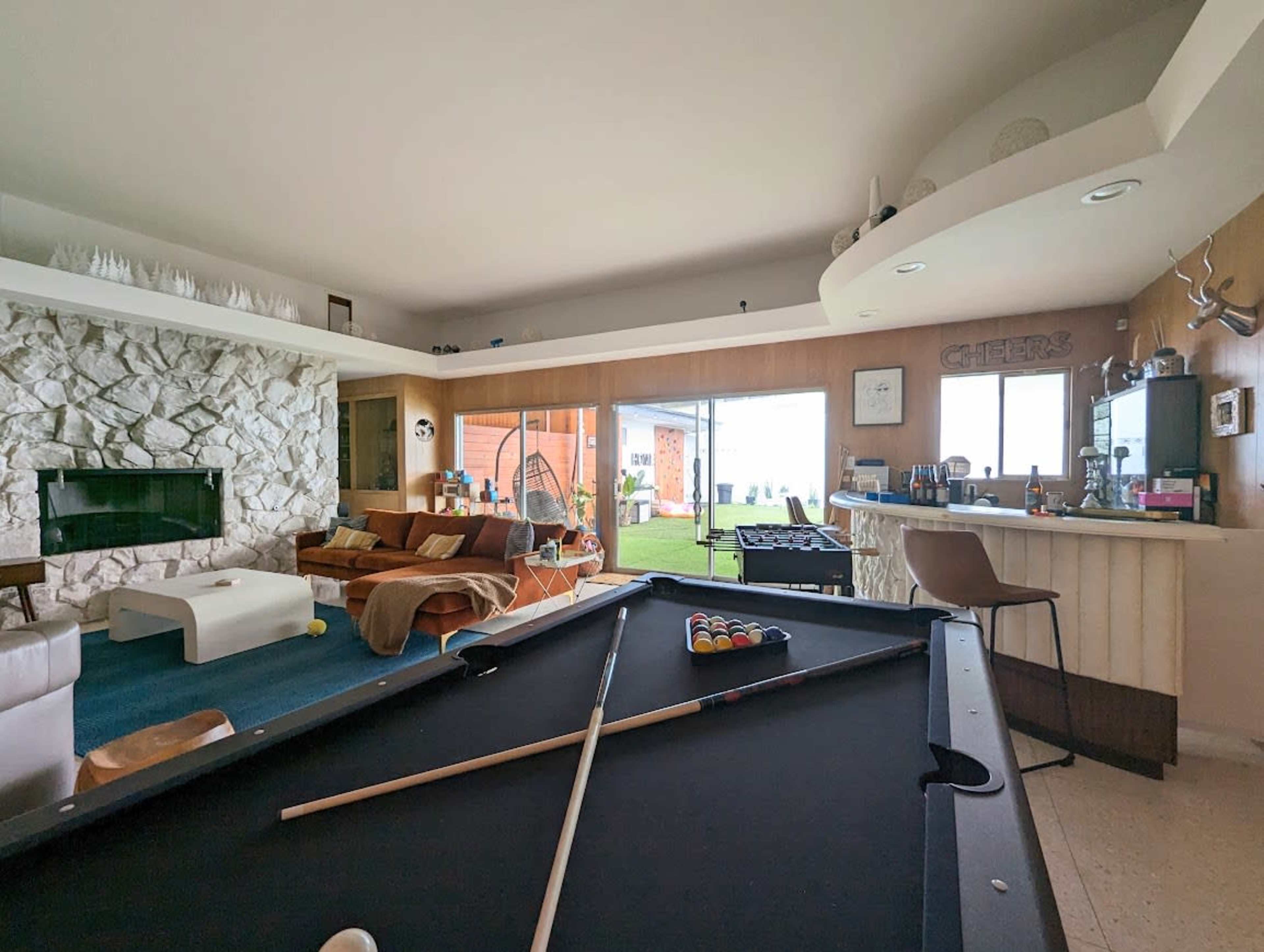 A spacious living area featuring a billiards table in the foreground, a stone fireplace, and a bar with large windows overlooking a grassy outdoor space.