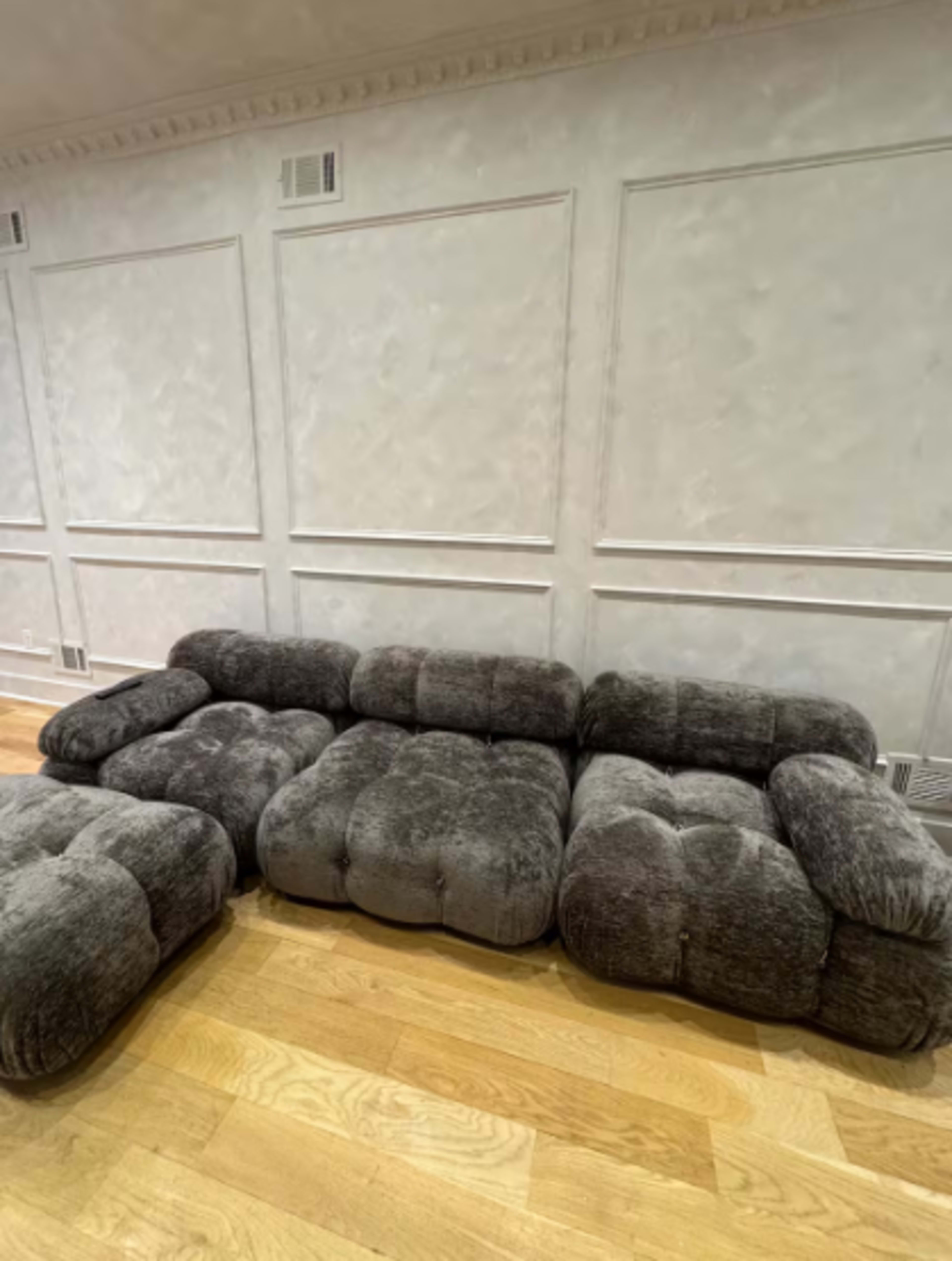 A gray, plush sectional sofa with a rounded design is positioned against a textured white wall on a wooden floor.