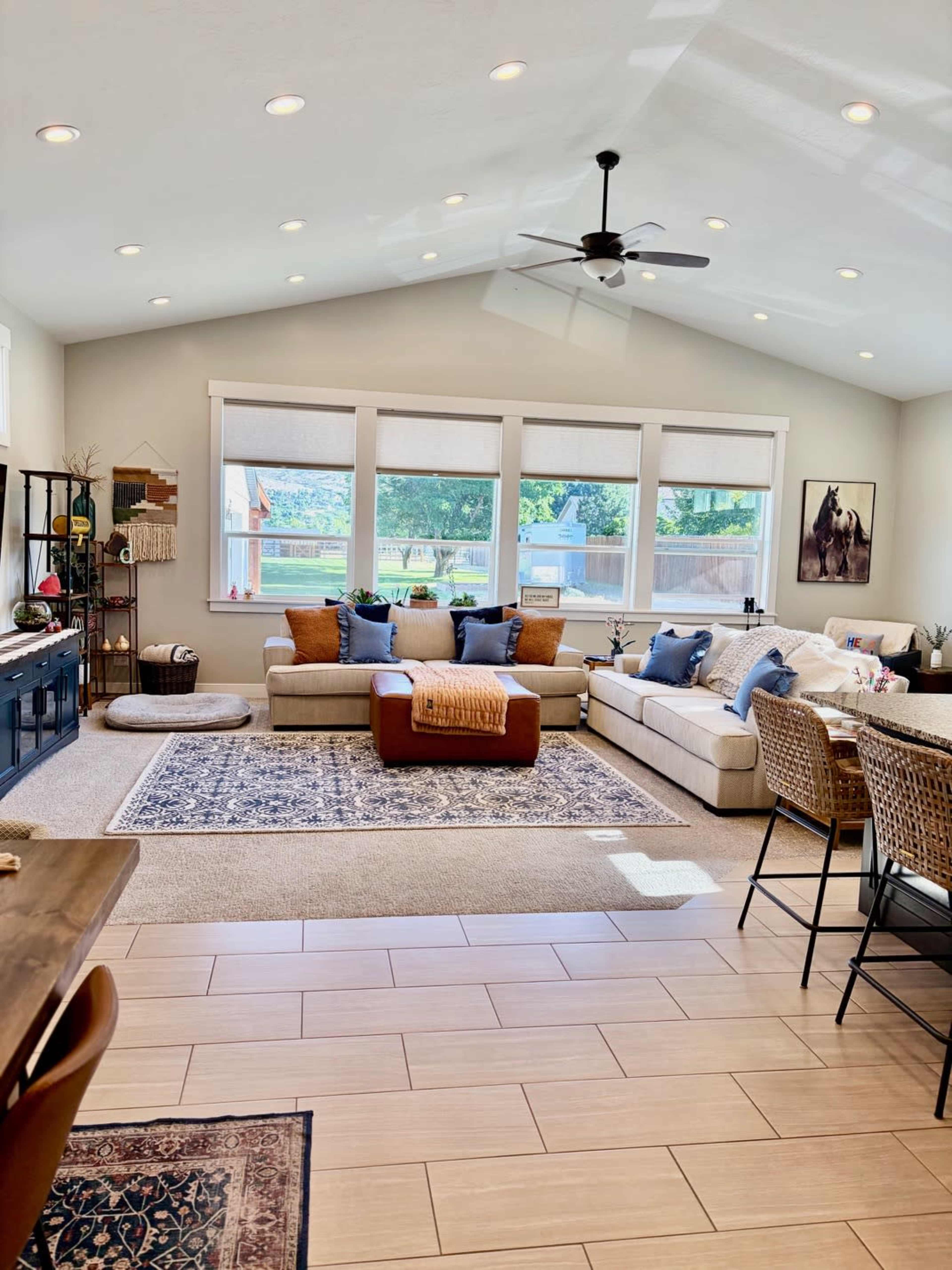 A bright living room features a large sectional sofa, a coffee table, and a dining area with wicker chairs, all arranged around a decorative area rug.