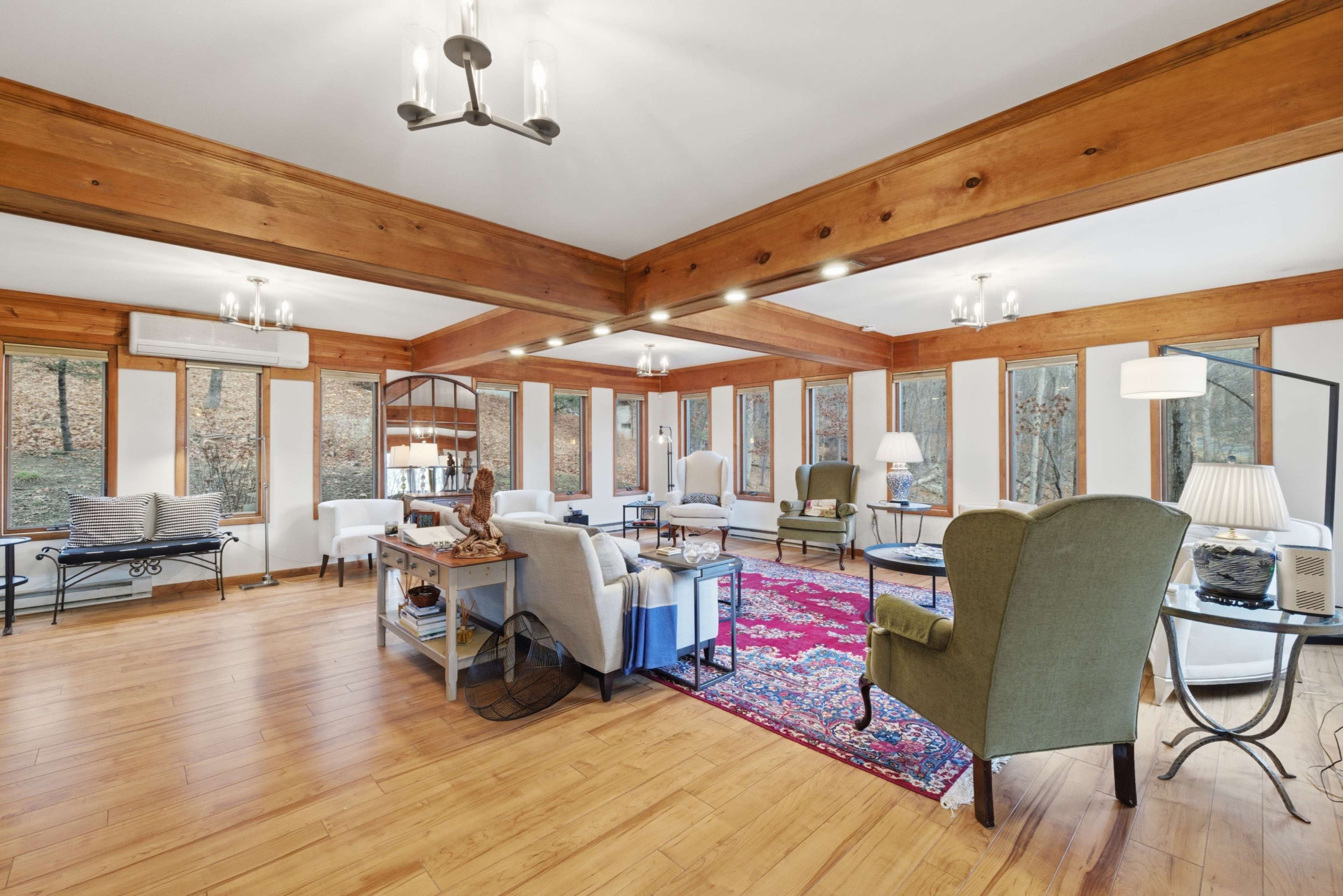 The spacious room features large windows, wooden beams, and a mix of seating arrangements including armchairs and a rug on the floor.