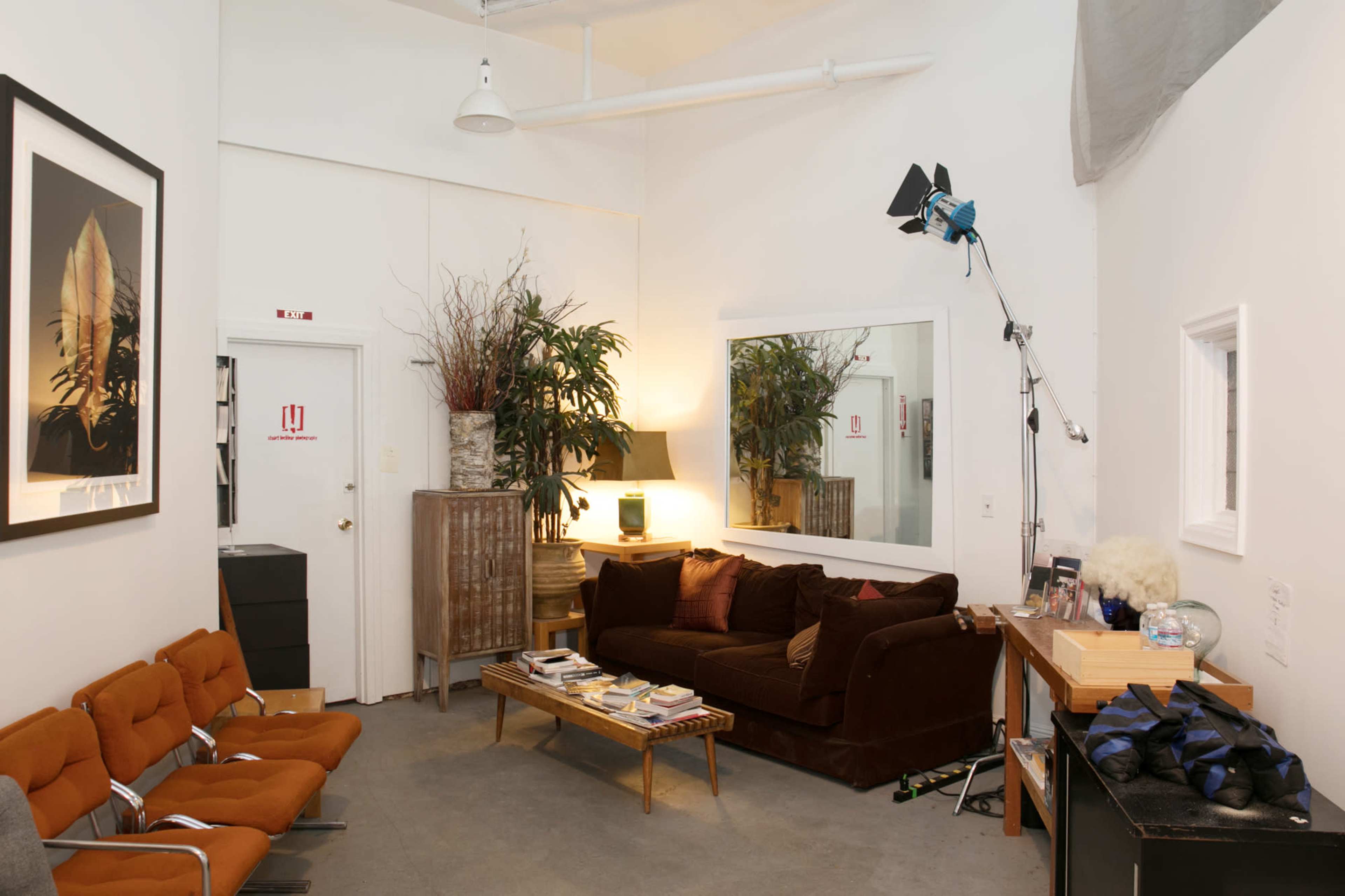 A small, well-lit room with a brown sofa, a coffee table stacked with magazines, potted plants, and a large mirror on the wall.
