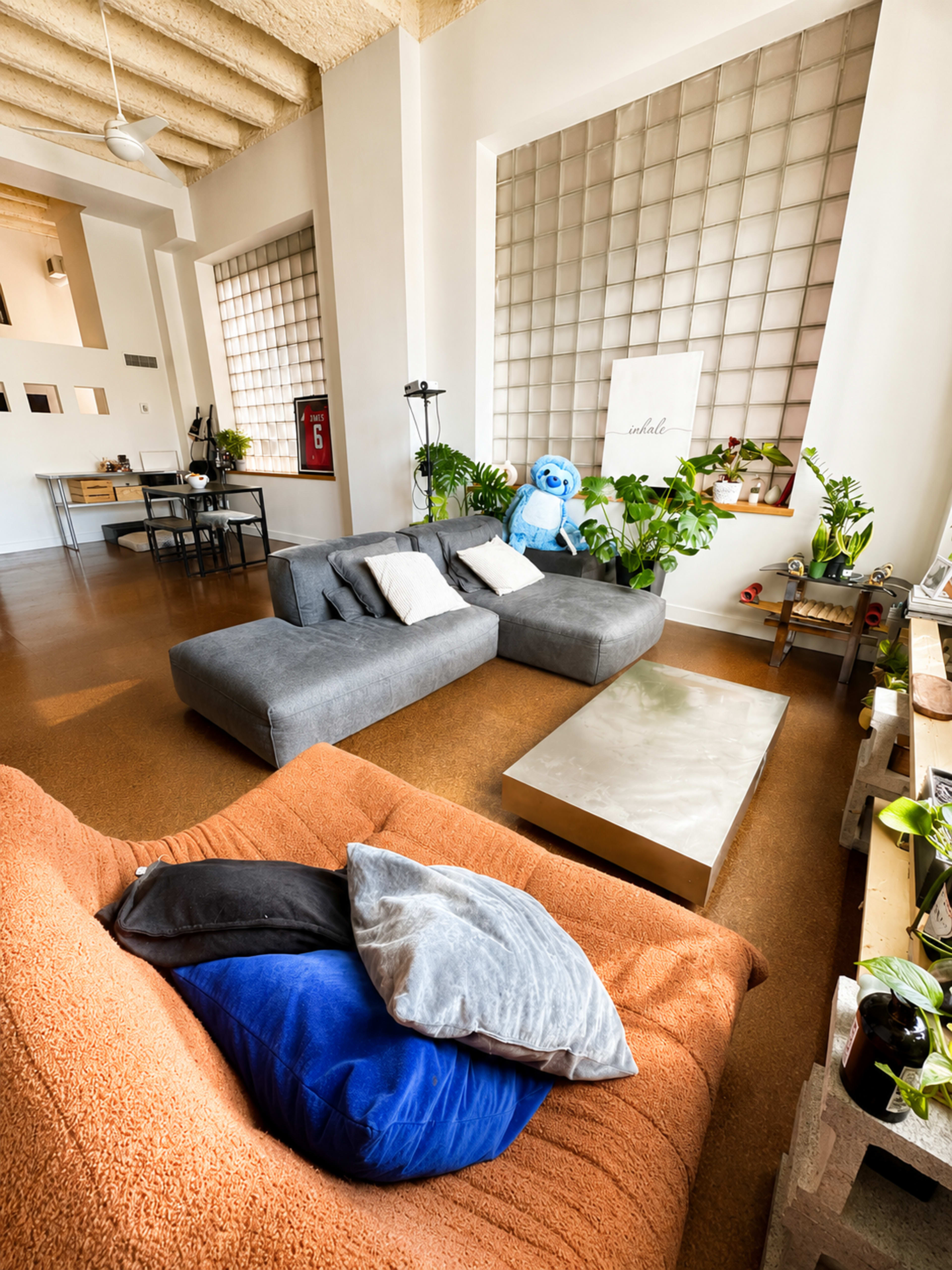 A cozy living room features a gray sectional sofa, colorful cushions, a metallic coffee table, and large windows with greenery.