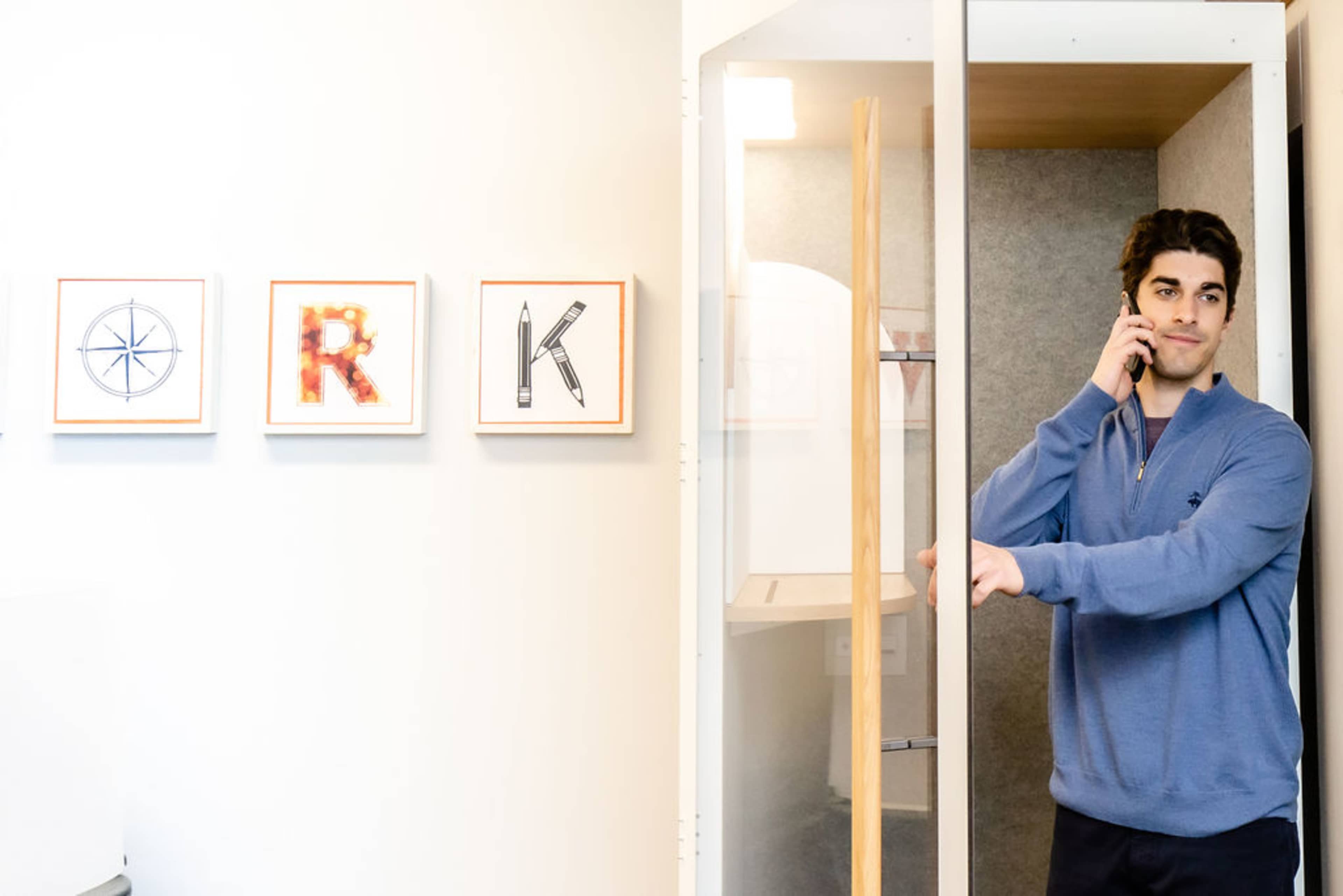 A man stands in an office corridor, holding a phone to his ear while appearing to exit a glass phone booth beside framed letters that spell "WORK."