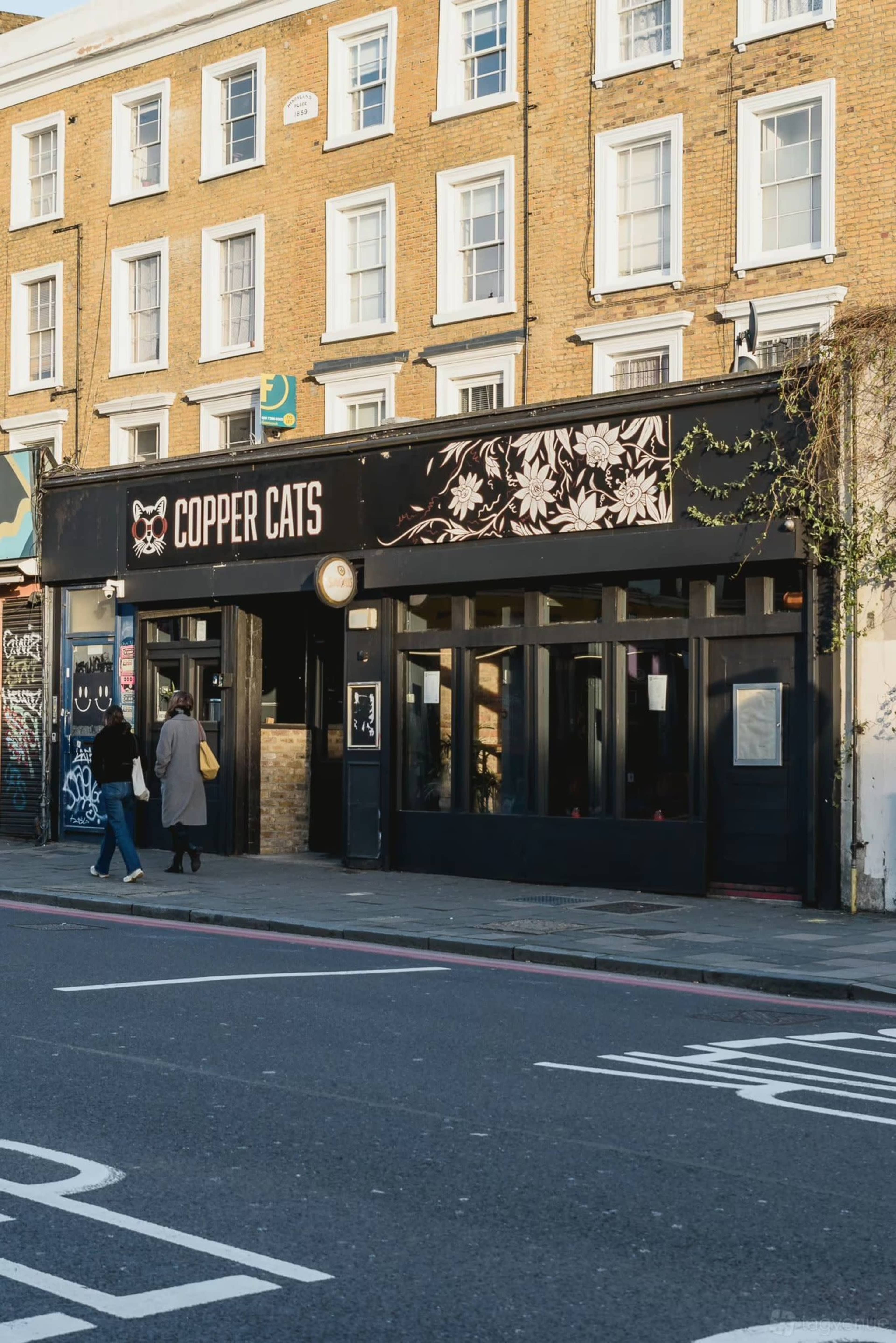 The image shows a bar named "Copper Cats" with a black exterior and floral designs, located on a street with pedestrians walking by.