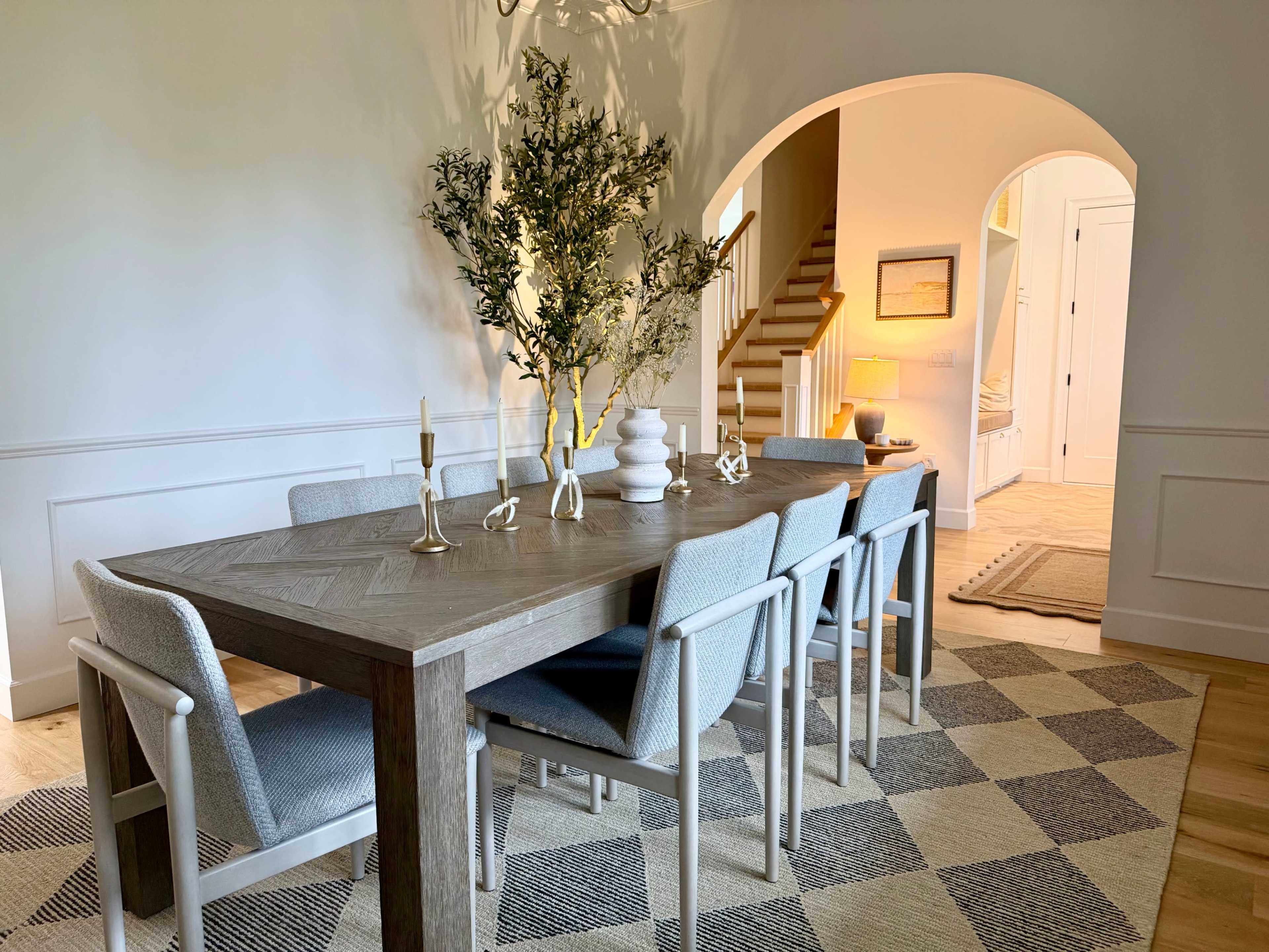 A dining room features a wooden table with gray chairs, a central floral arrangement, and an archway leading to a staircase and a softly lit hallway.