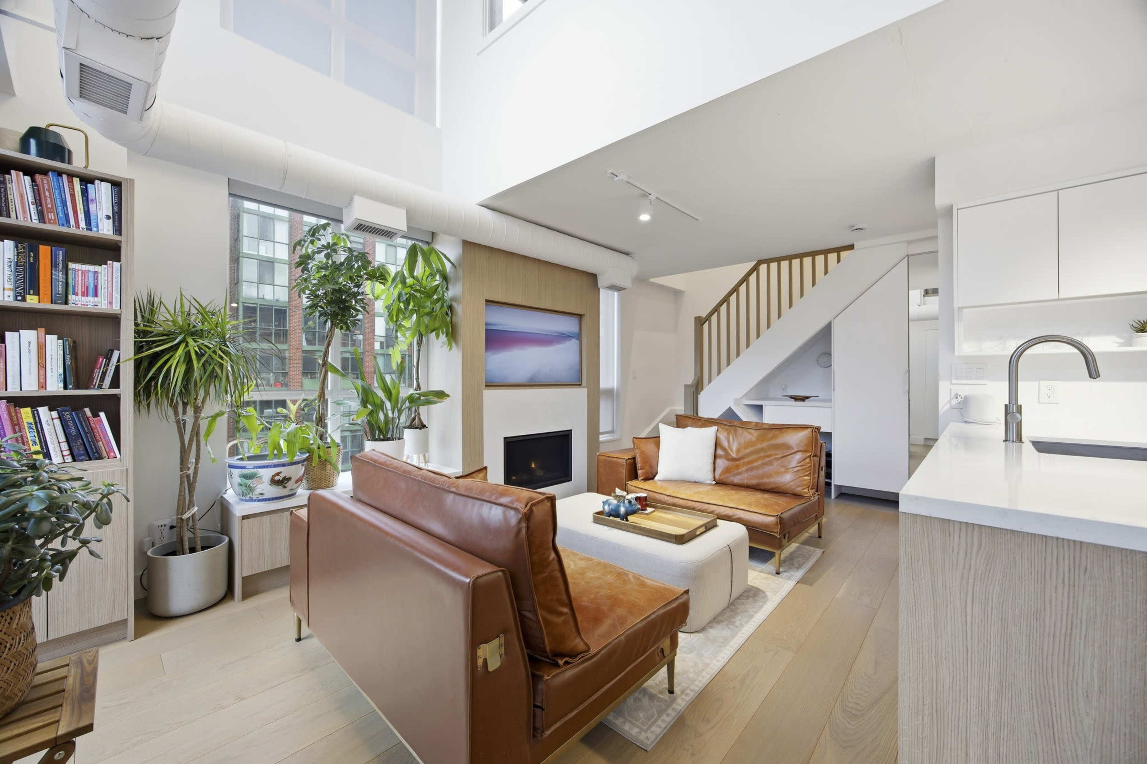 The image shows a modern living room featuring leather sofas, a coffee table, a plant shelf, and an open kitchen area with light wood finishes and a small staircase.