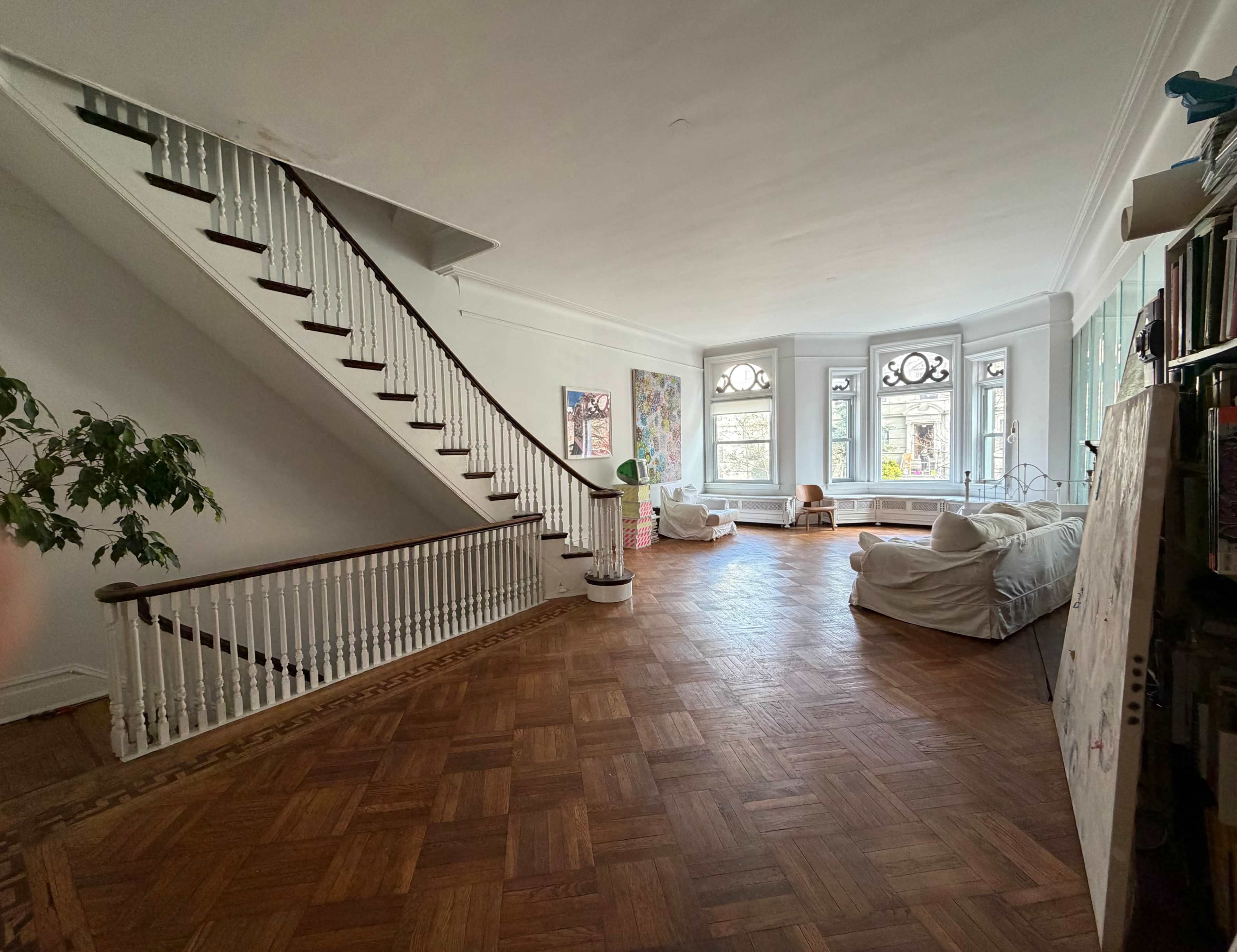 The image shows a spacious room with polished wooden floors, a staircase on one side, and large windows allowing natural light to fill the space.