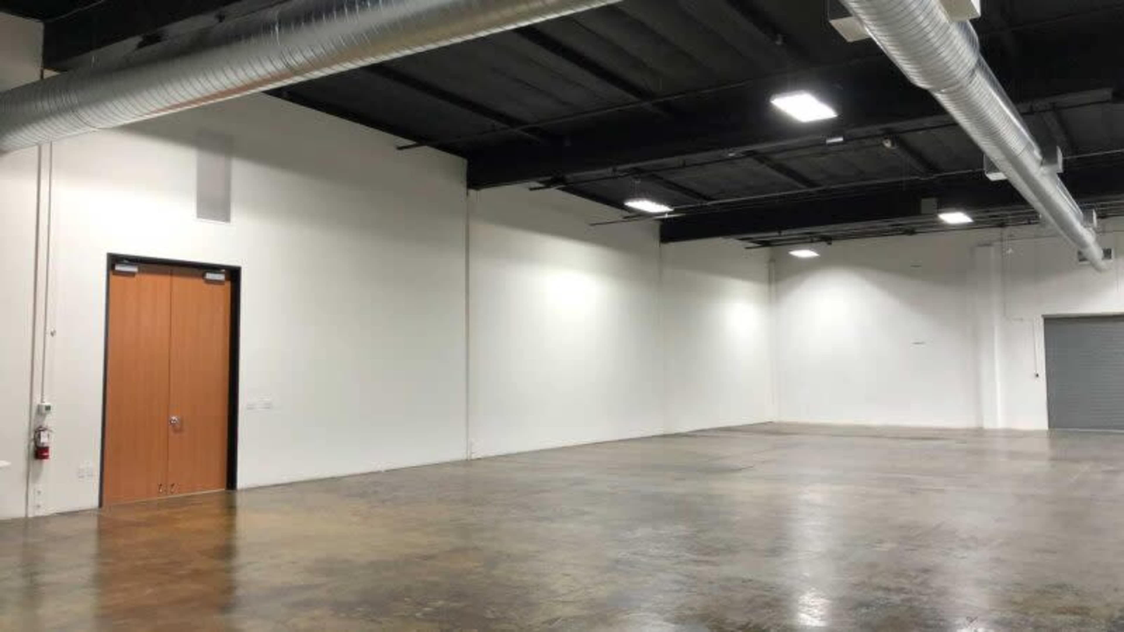 A large, empty industrial space with white walls and a concrete floor, featuring a wooden door and a roll-up door on opposite walls.
