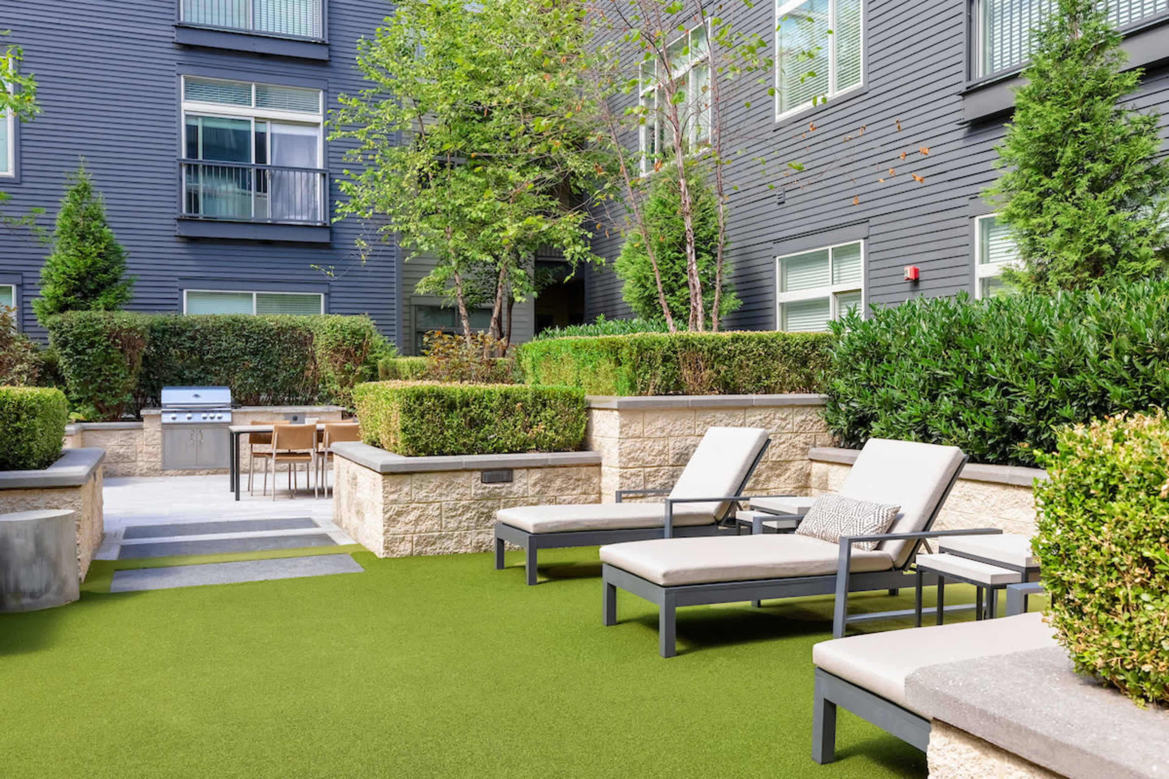 A landscaped courtyard features lounge chairs, a barbecue grill, and a dining area surrounded by neatly trimmed hedges and trees.
