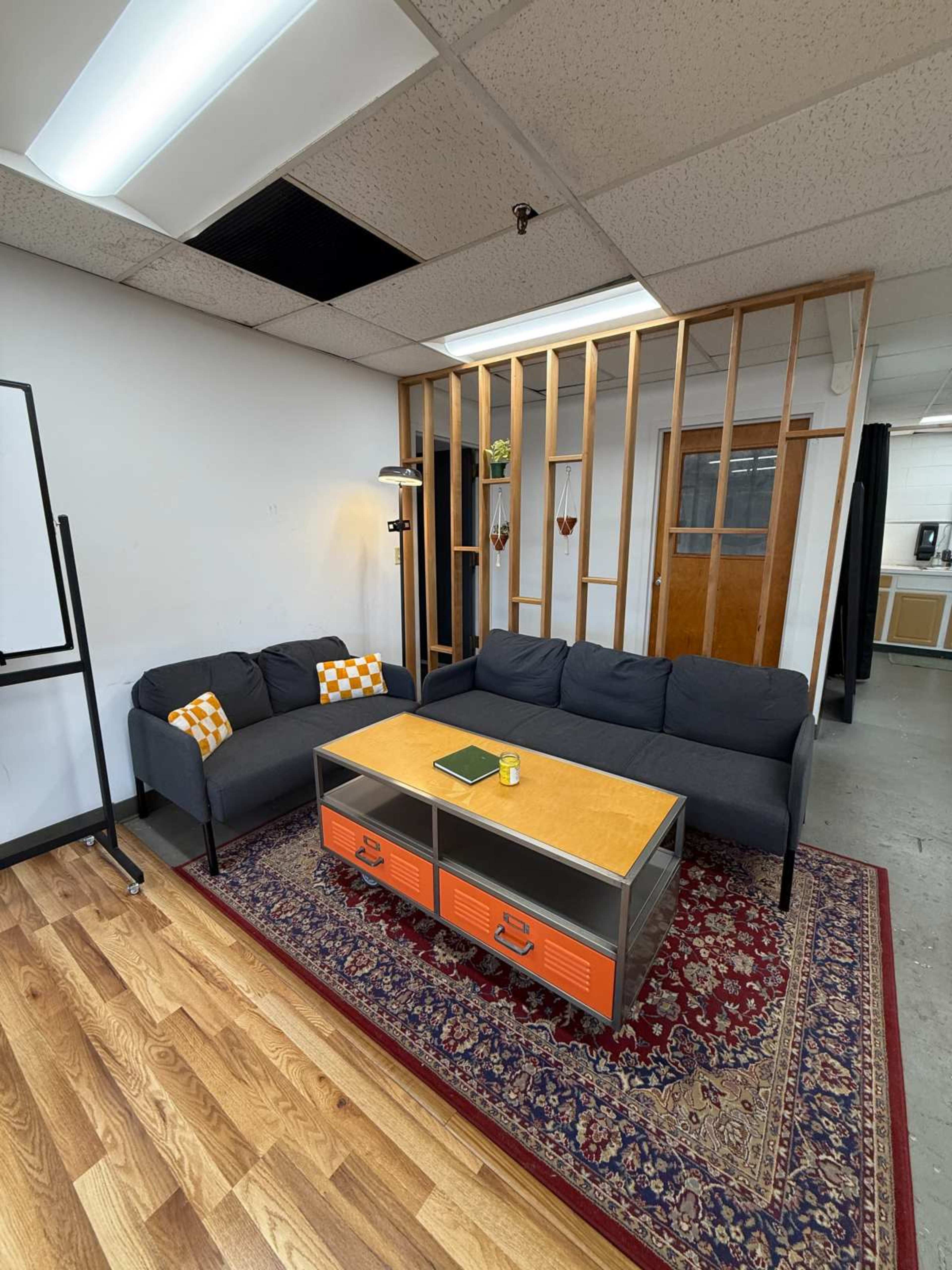 The image shows a modern office space featuring a gray sofa with an orange and white checkered cushion, a coffee table with orange drawers, a decorative rug, and a wooden partition.