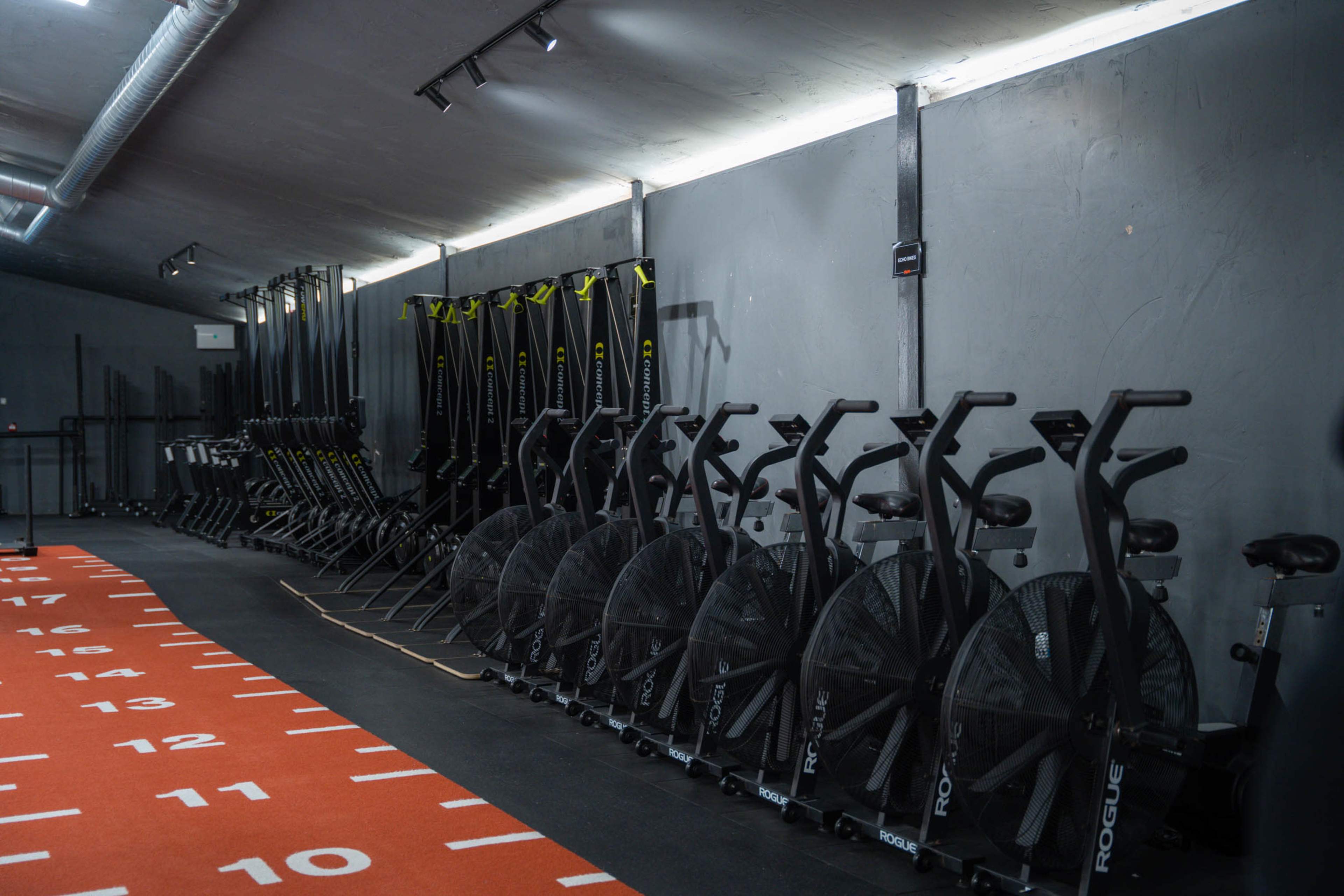 A fitness gym with rows of exercise bikes and rowing machines lined against a gray wall, and a track marked with numbers on the floor.
