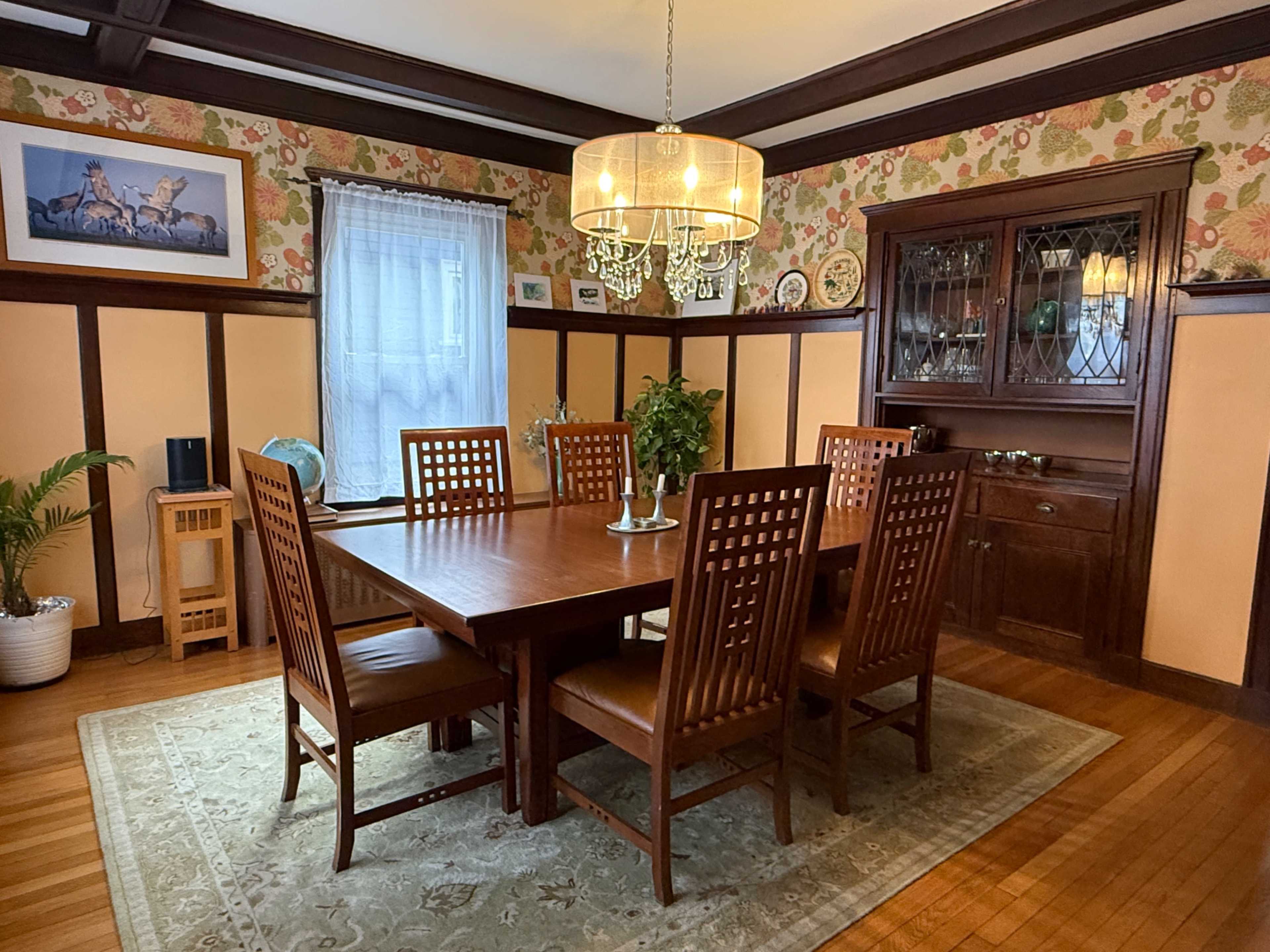 The image shows a dining room with a large wooden table surrounded by eight chairs, a chandelier overhead, and floral-patterned wallpaper.