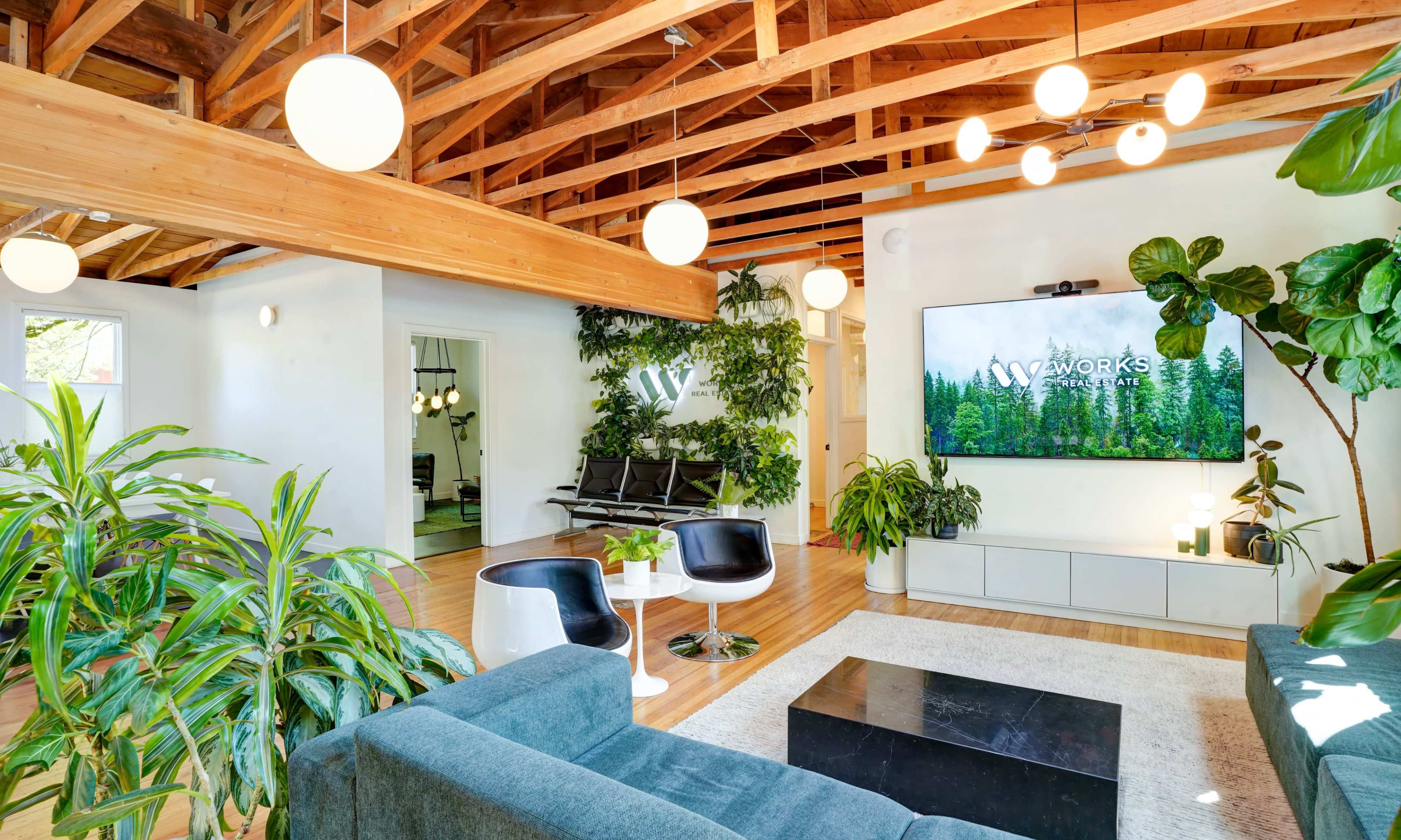 The image shows a modern living space with a wooden-beamed ceiling, green plants, a large television mounted on the wall, and stylish furniture arranged around a coffee table.