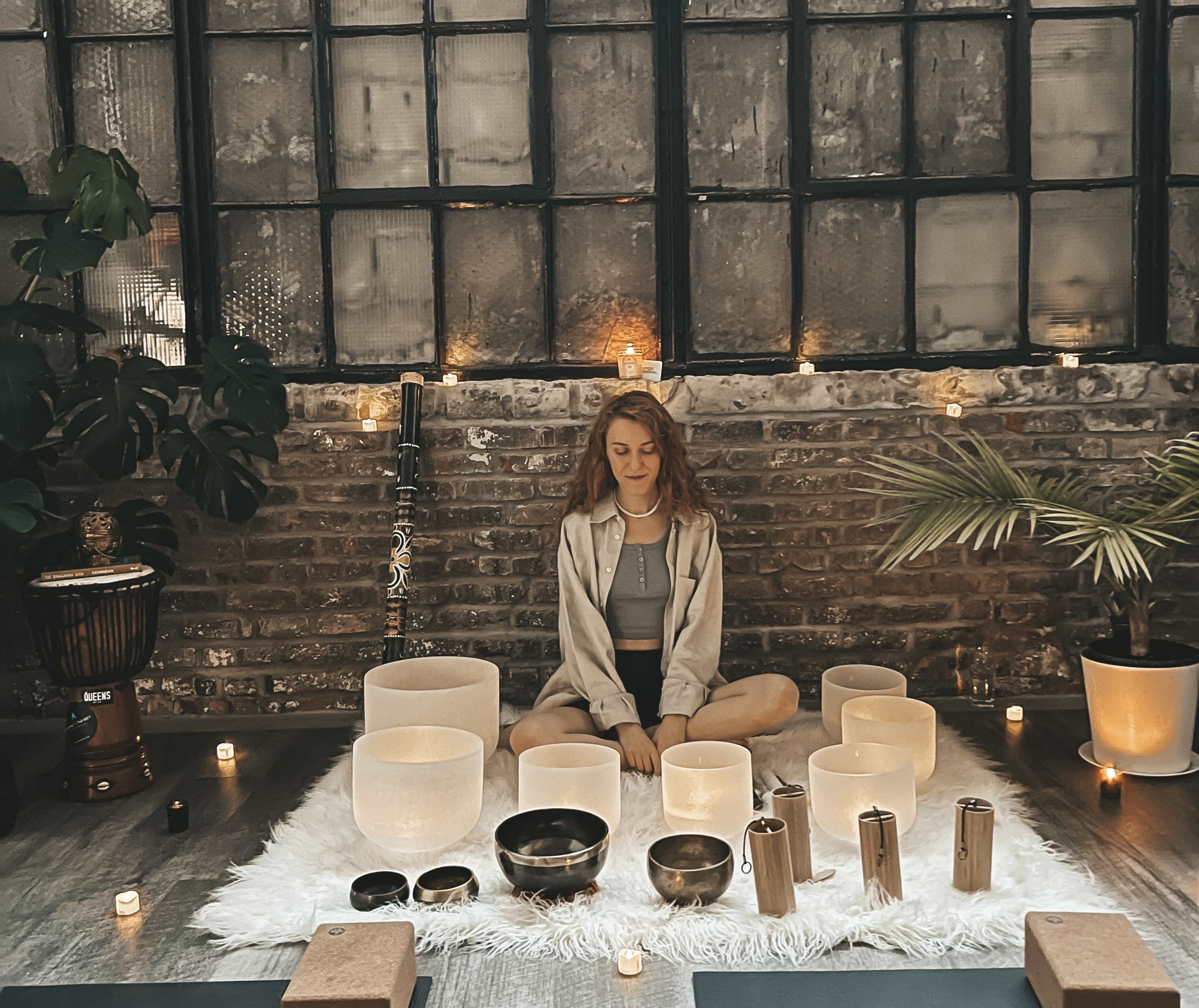 A person sits cross-legged on a white rug surrounded by various sound bowls, candles, and plants in a room with large windows.