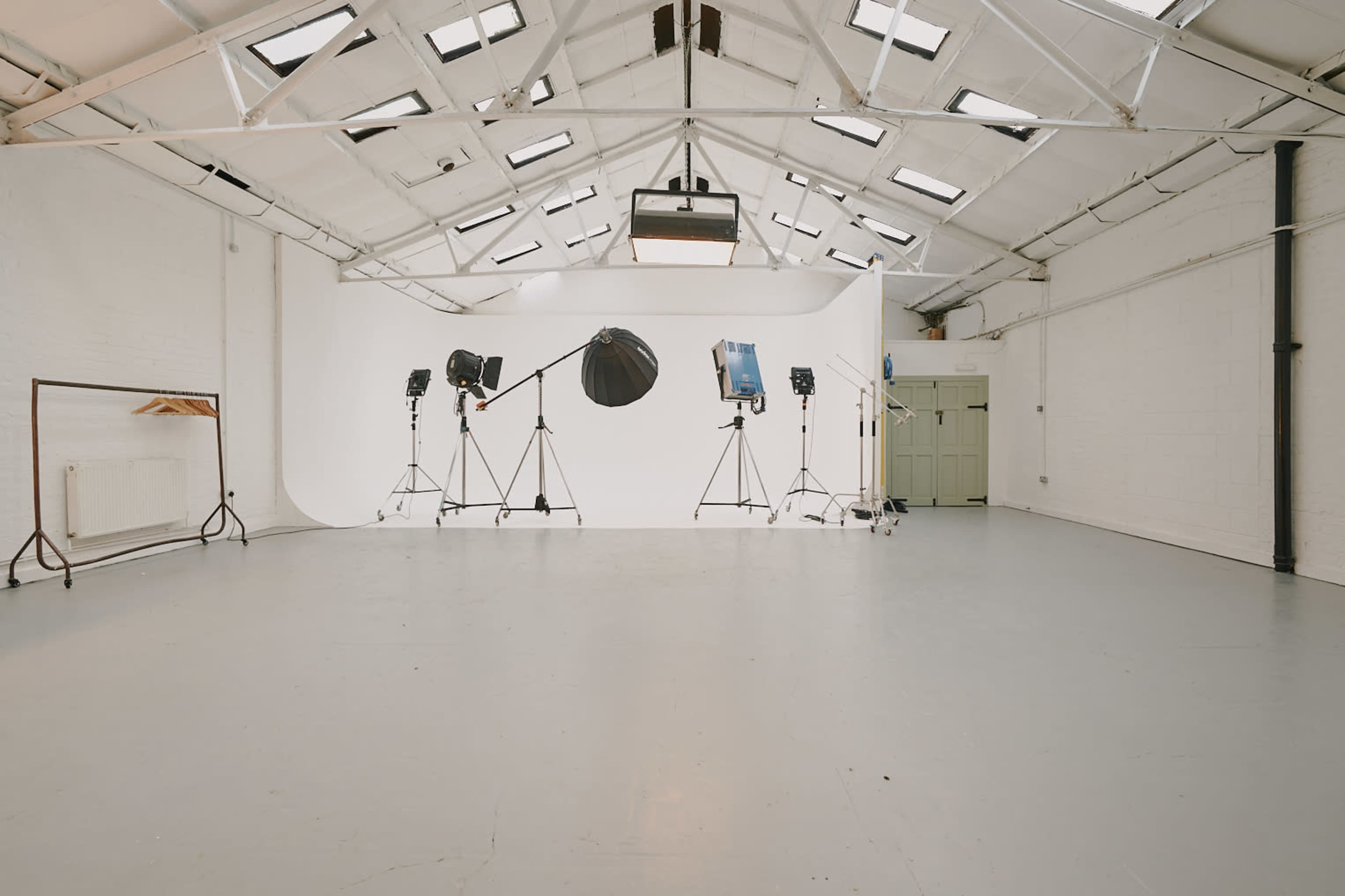 A spacious photography studio featuring a white backdrop, multiple lighting equipment on stands, and a wooden rack against a wall.