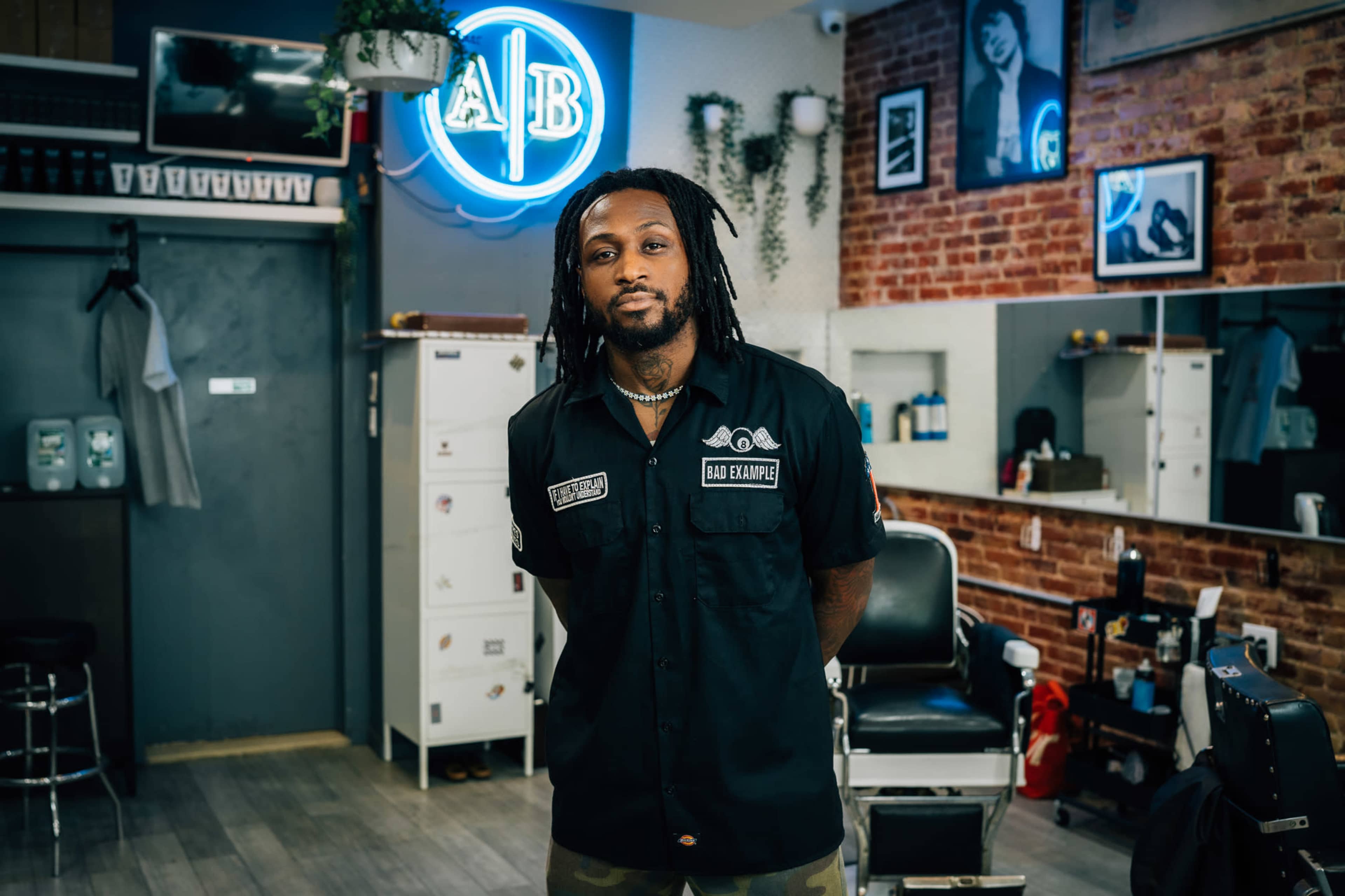 A barber stands in a well-lit barbershop with a neon sign and brick walls.