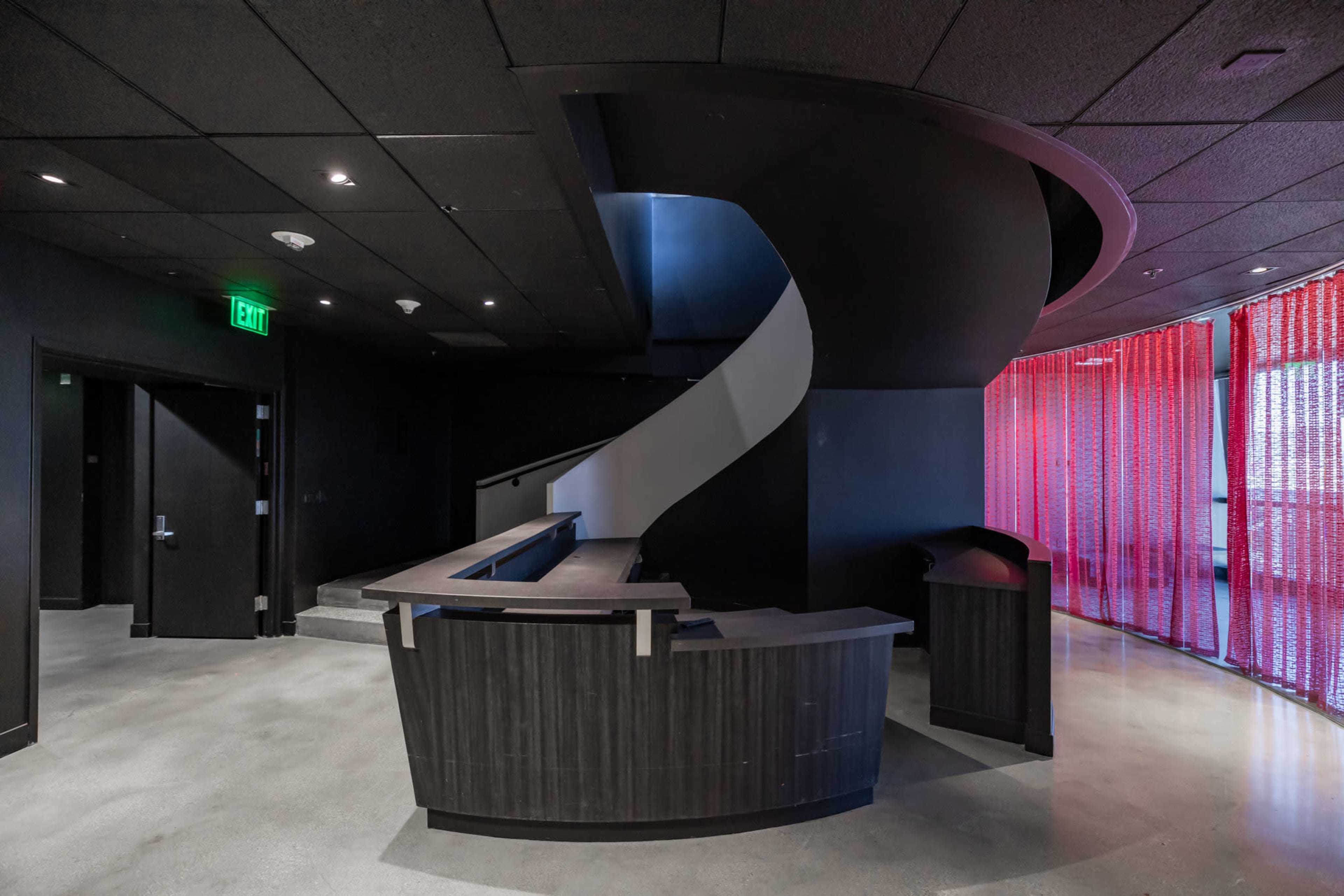 The image shows a modern interior with a curved staircase, a dark reception desk, and a wall featuring red translucent panels.