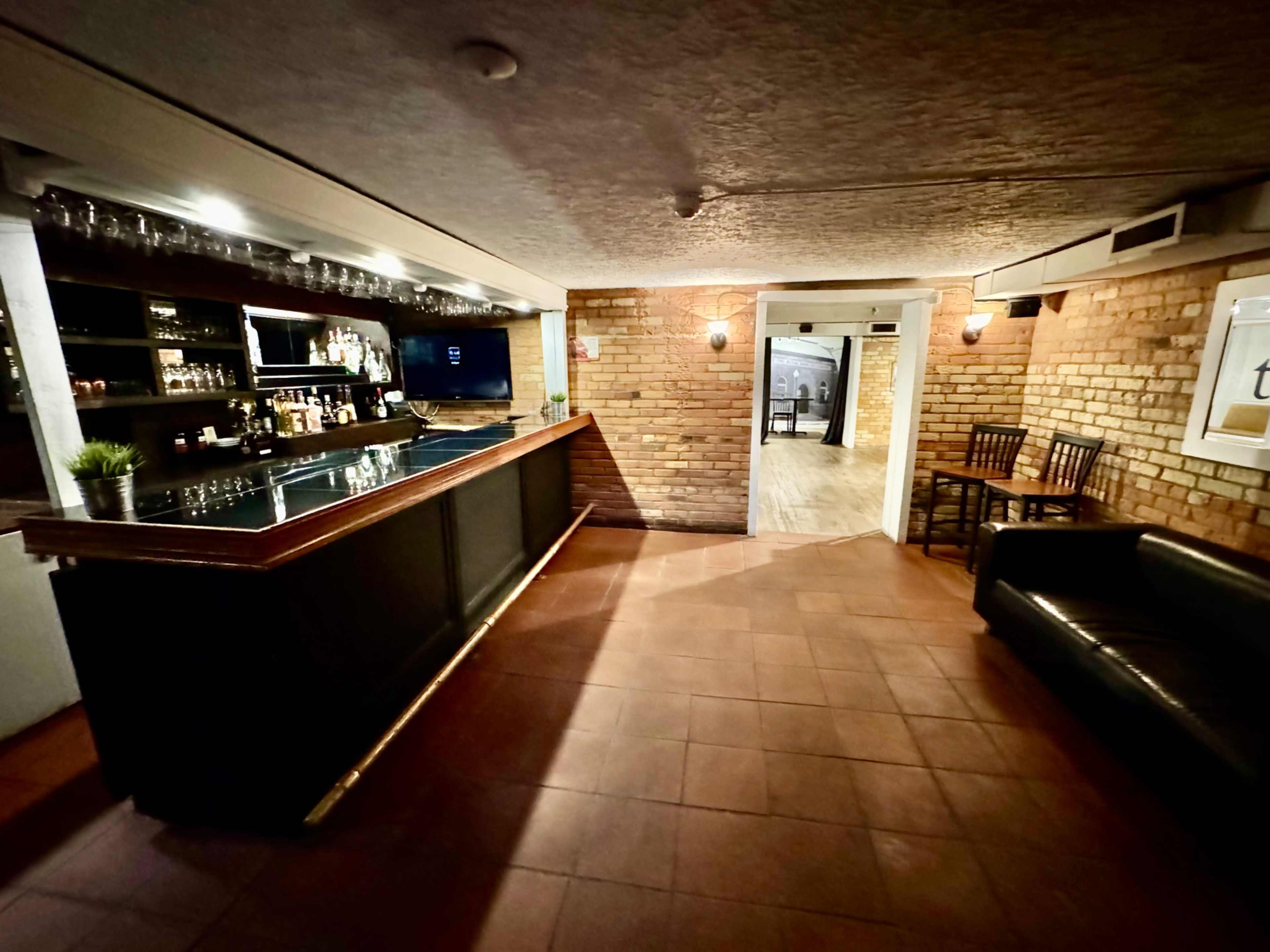 A dimly lit bar area with a wooden counter, shelves of bottles, and a doorway leading to another room.
