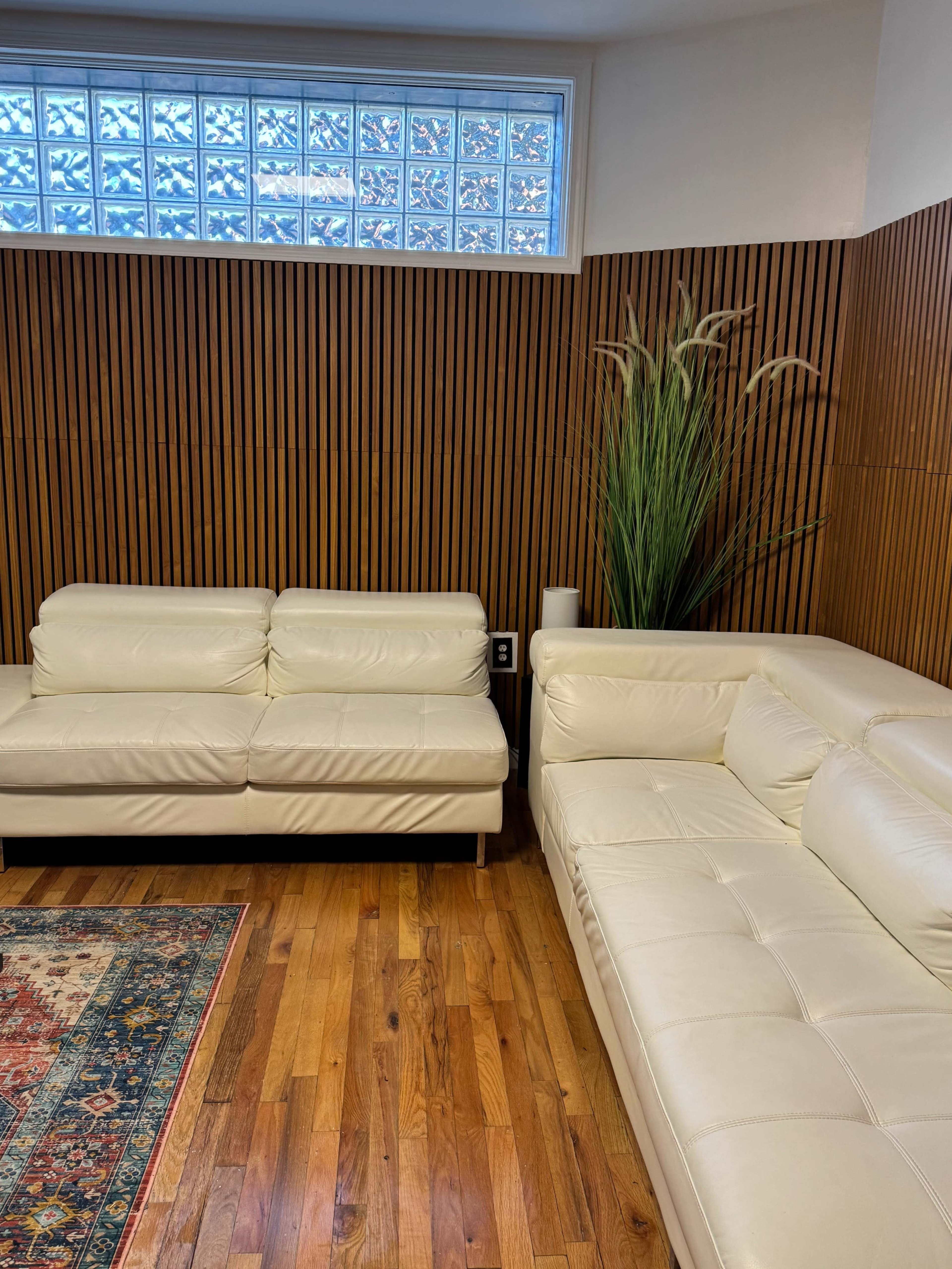 The image shows a living room with two white leather sofas, a colorful area rug on wooden flooring, and a wall featuring wooden paneling and a glass block window.