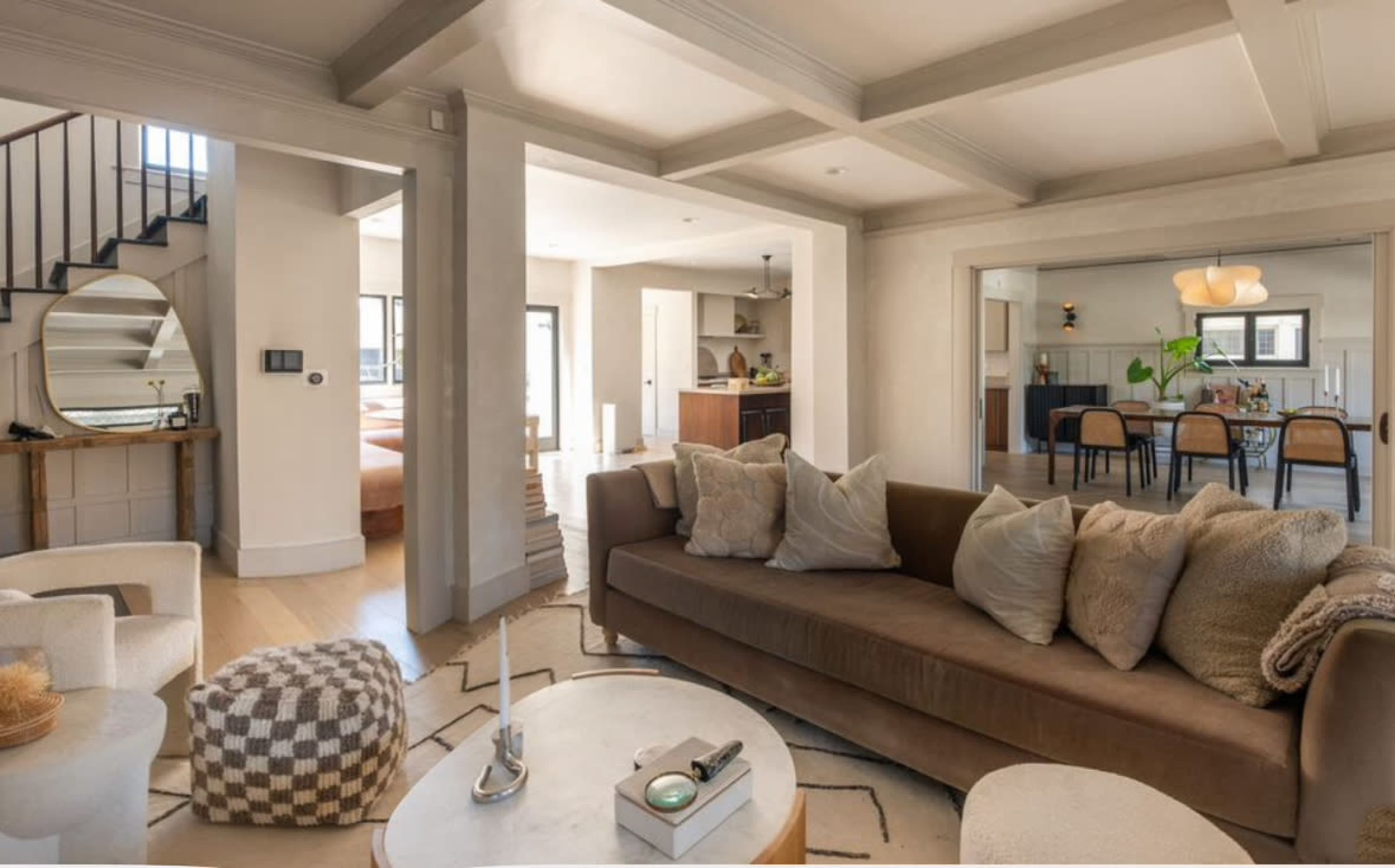 The image shows a modern interior living space featuring a brown sofa with cushions, a round coffee table, and an open layout connecting to a dining area and staircase.