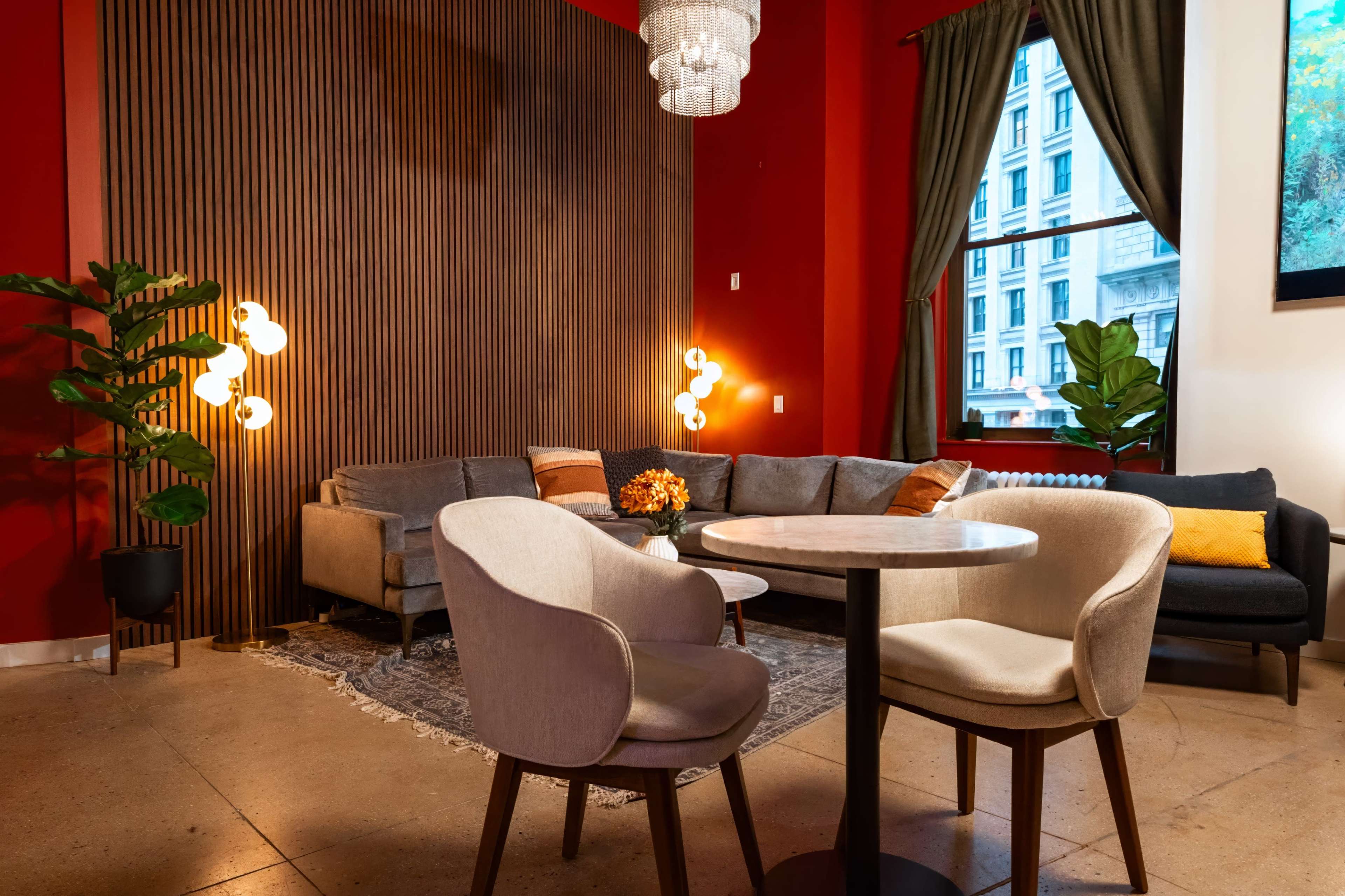 The image shows a cozy indoor seating area featuring a gray sofa, a round table with two upholstered chairs, and decorative plants against a backdrop of red and wooden paneling.