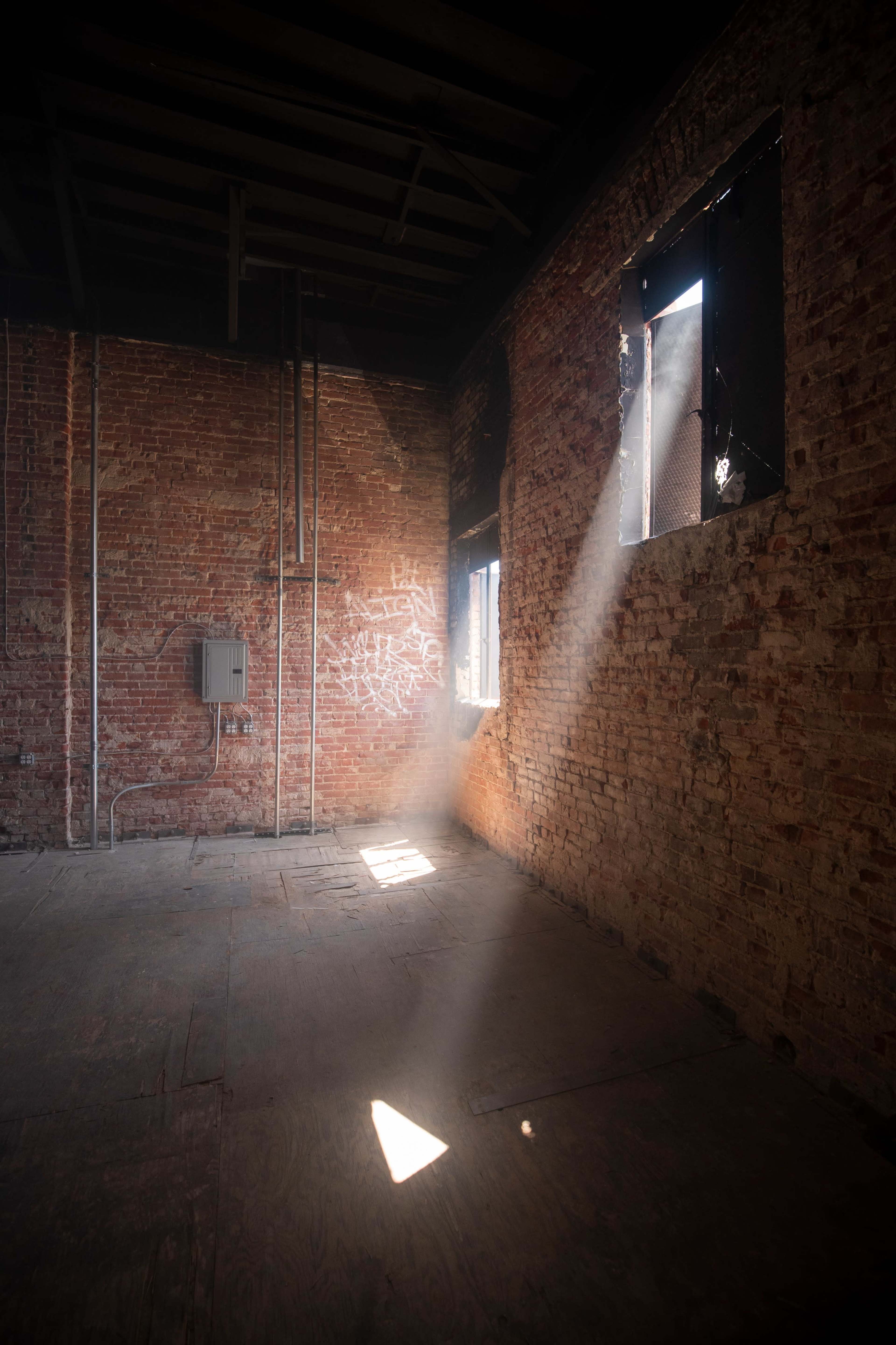 Light streams through a window, illuminating the dust in a dimly lit room with exposed brick walls and wooden flooring.