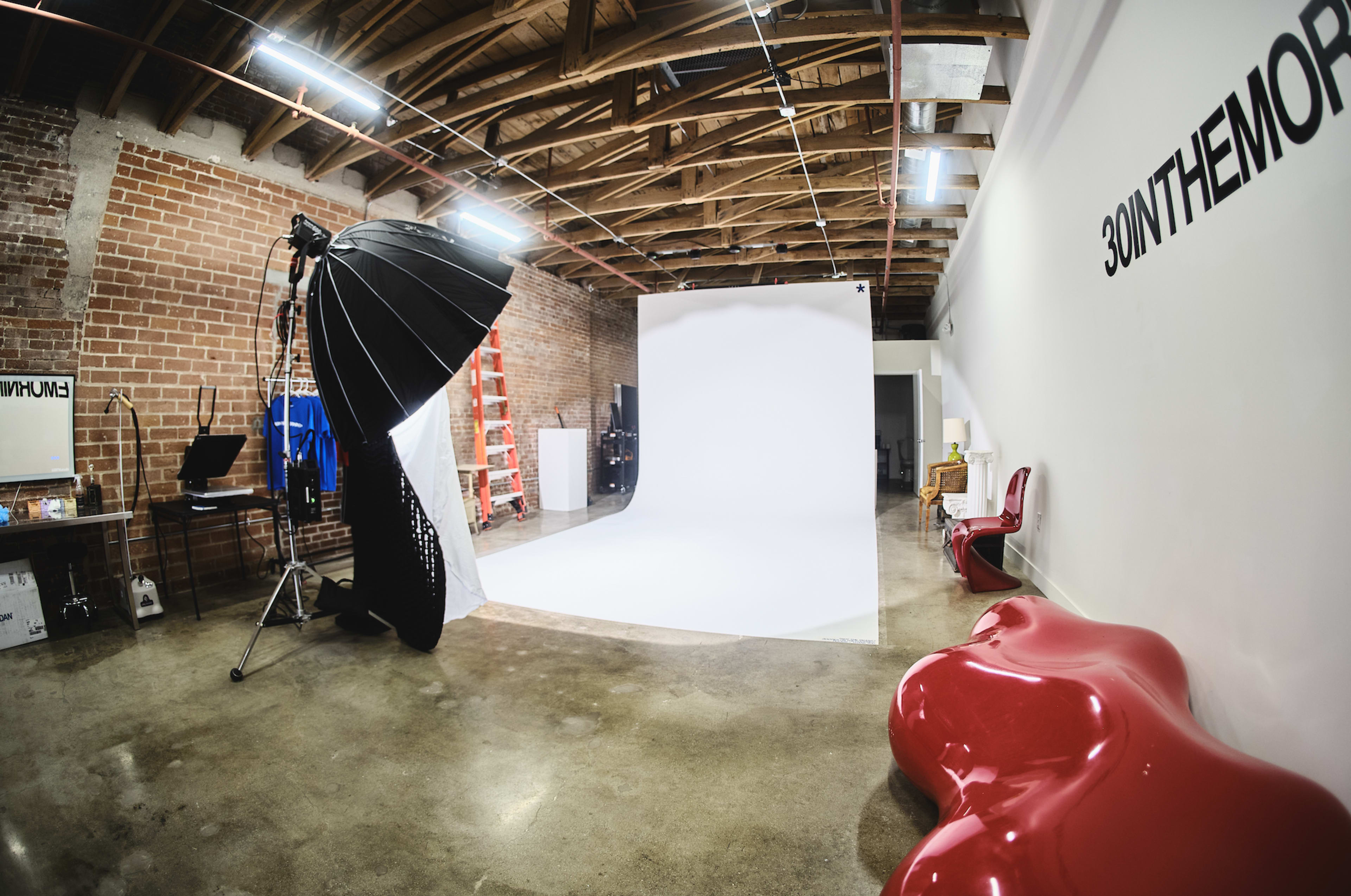 The image shows a spacious photography studio with a large white backdrop, a softbox light setup, and modern furniture against a brick wall.
