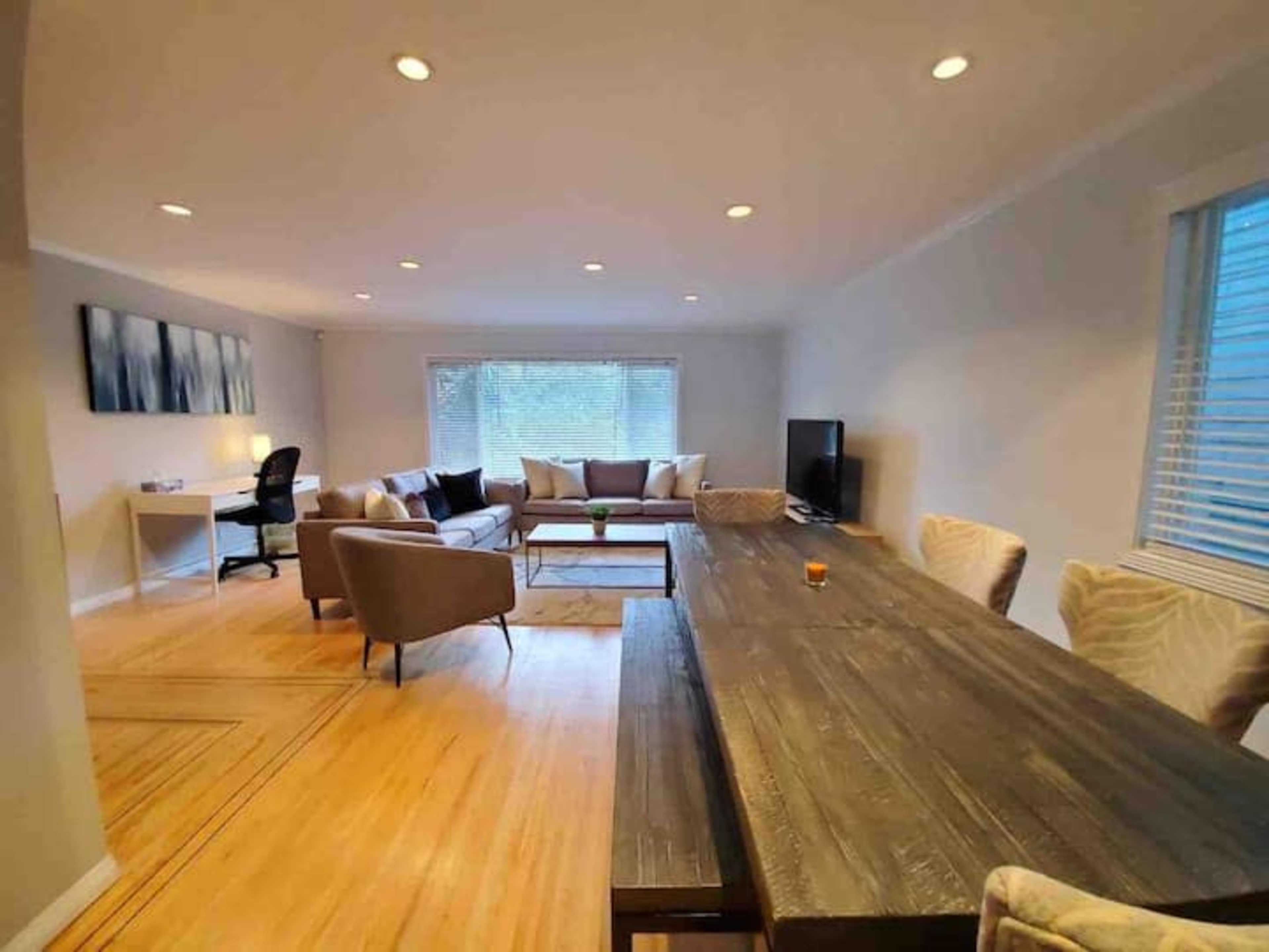 The image shows a modern living room with a wooden dining table, a sofa arrangement, a television, and a desk with a chair, all illuminated by recessed lighting.