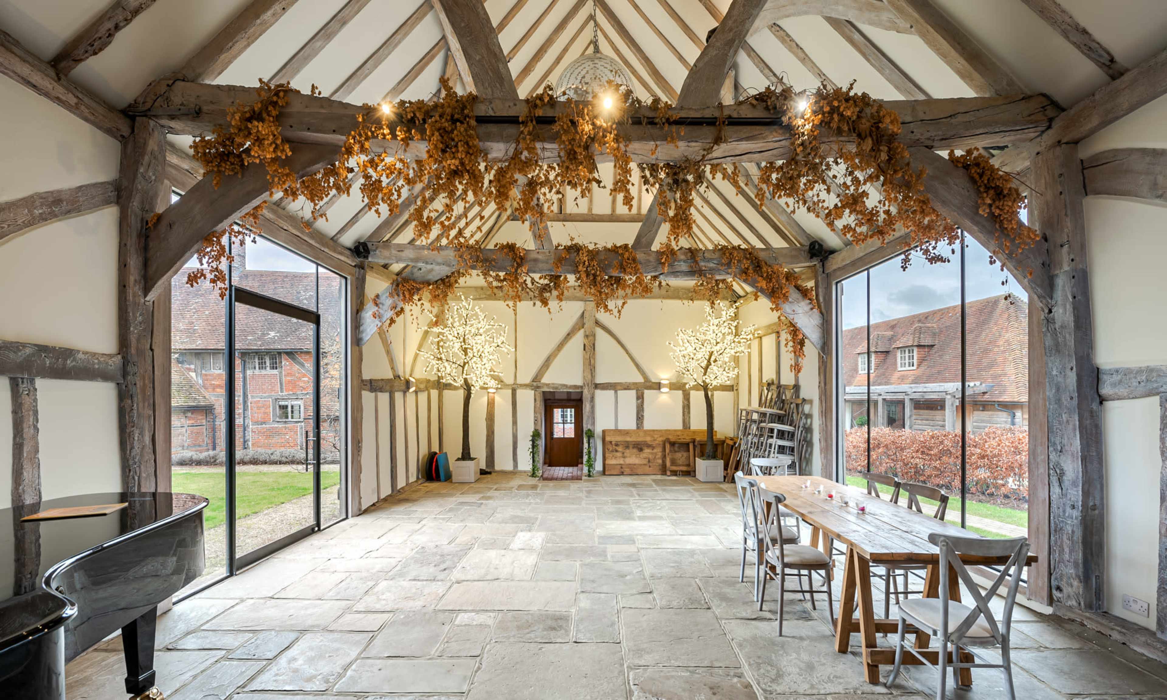 A spacious interior with exposed wooden beams, a stone floor, large glass doors, a dining table, a piano, and decorative branches hanging from the ceiling.
