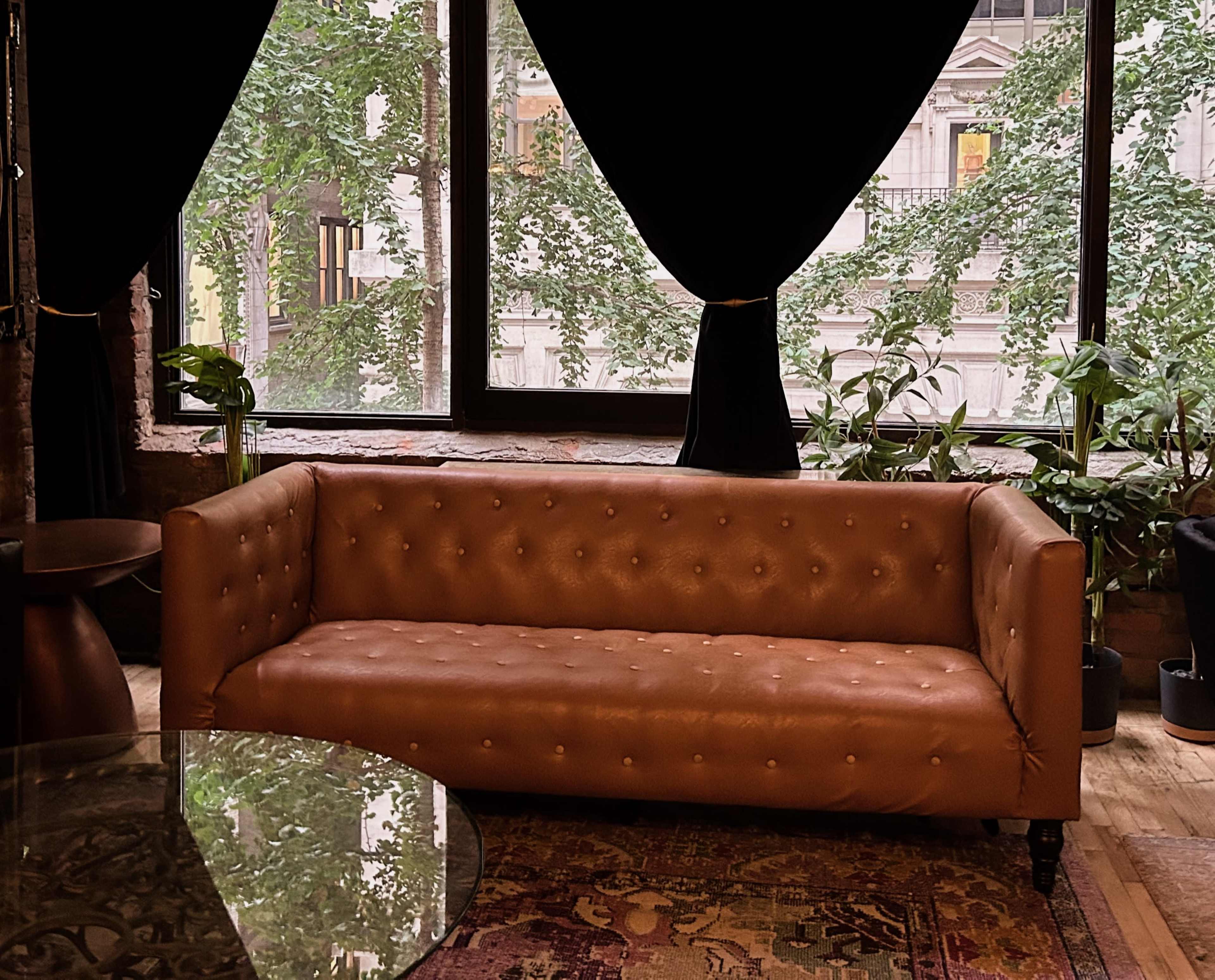 A brown leather sofa sits in front of large windows adorned with dark curtains, surrounded by indoor plants.