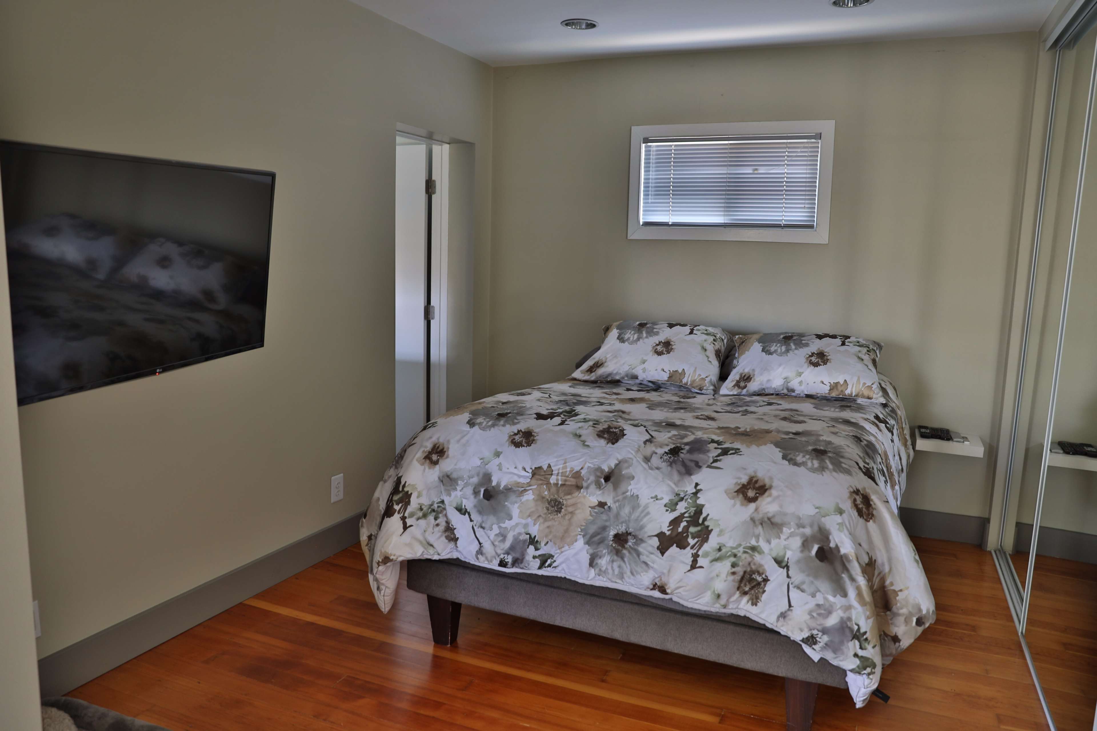 The image shows a simple bedroom with a large bed covered in a floral-patterned comforter, a flat-screen TV mounted on the wall, and a mirror reflecting the room's interior.