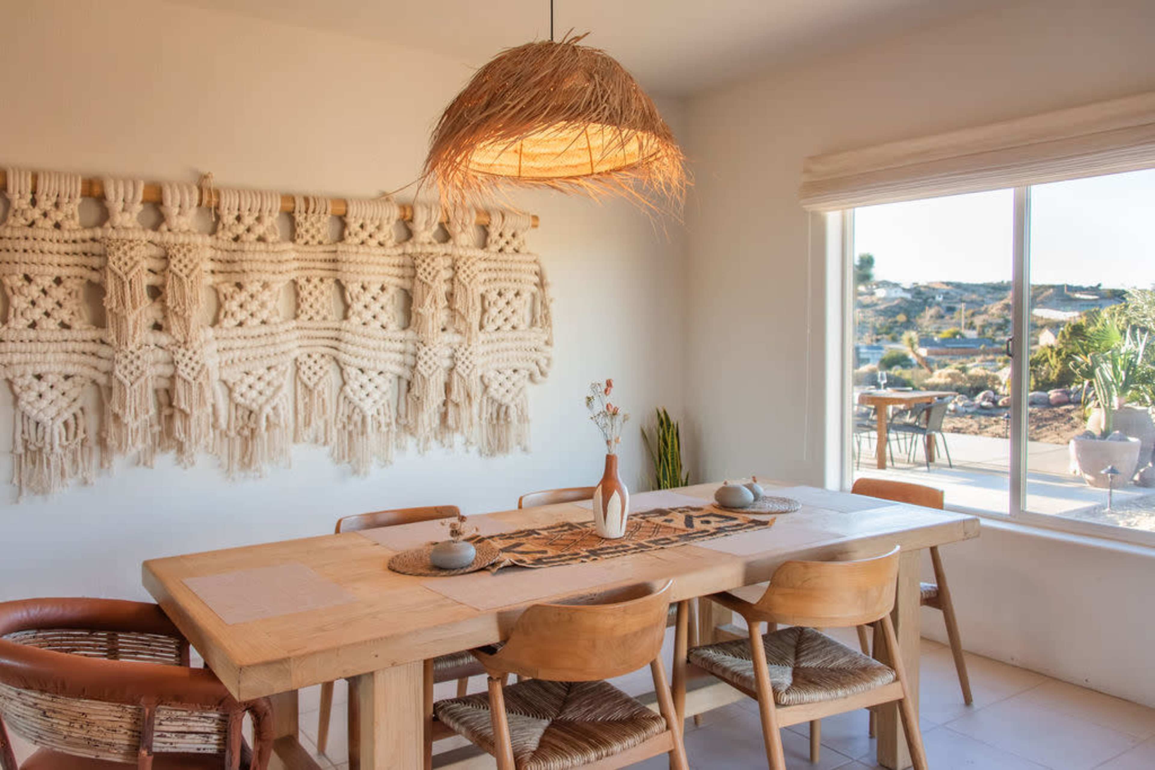 A wooden dining table with six chairs sits beneath a woven light fixture, next to a large window showcasing an outdoor view, while a macramé wall hanging adorns the wall behind it.