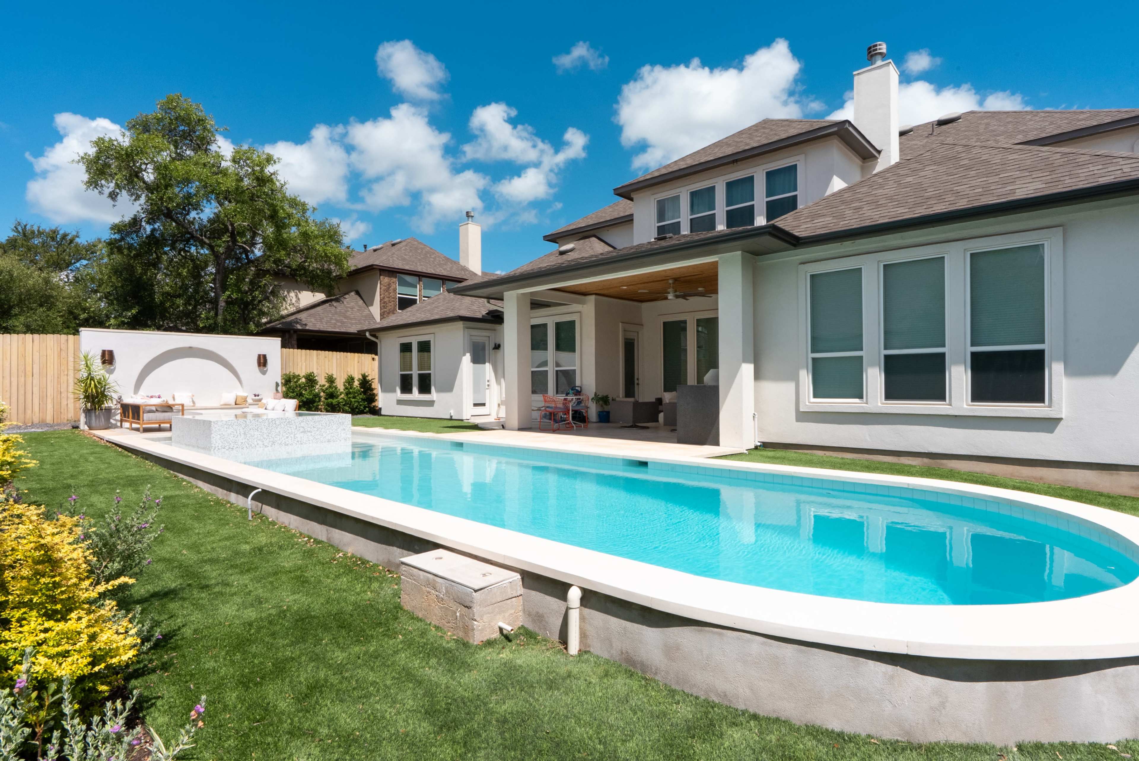 A spacious backyard features a rectangular swimming pool alongside a modern house with multiple windows and a covered patio area.