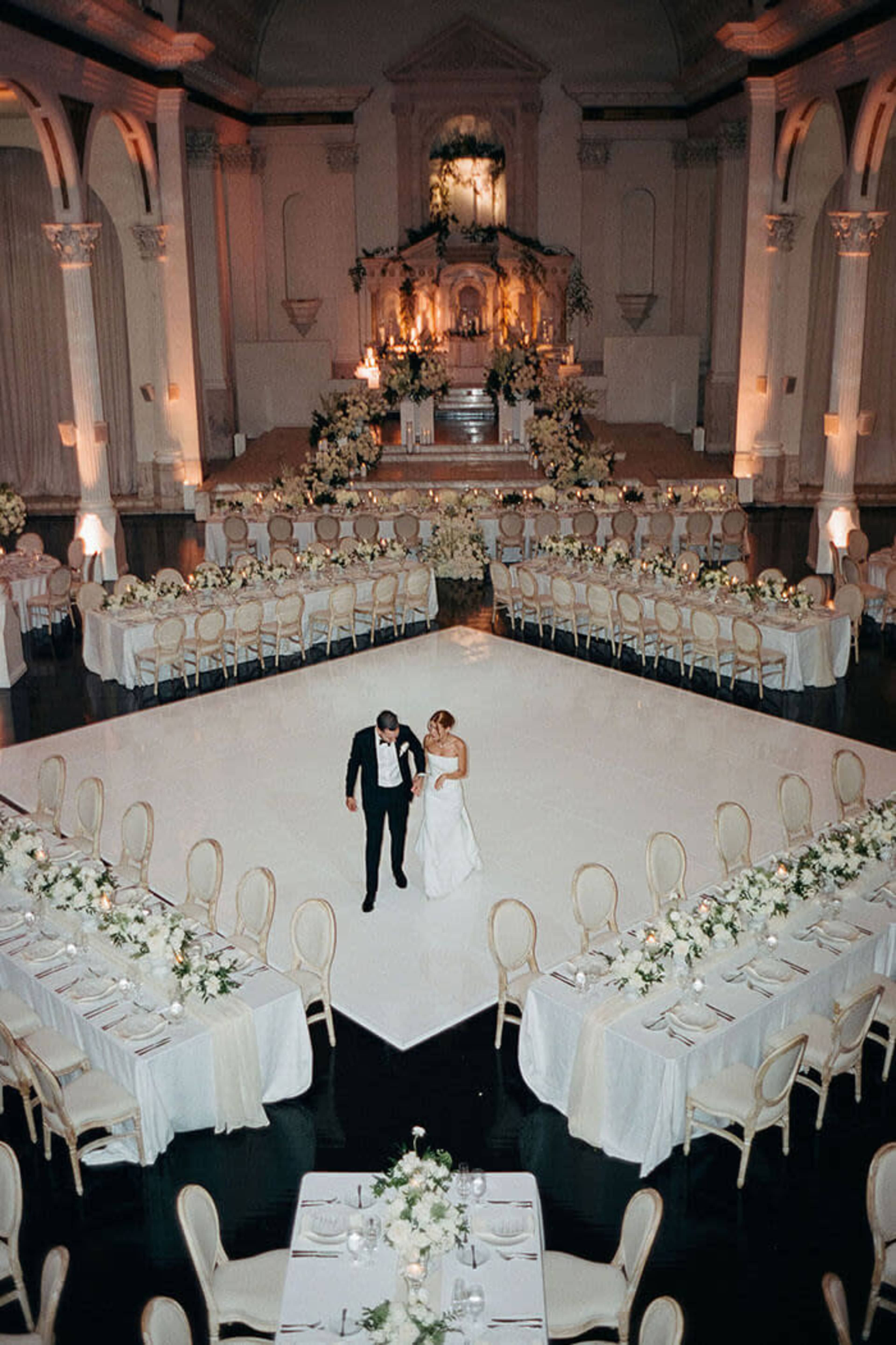A couple walks hand in hand on a white dance floor surrounded by elegantly set tables in a grand wedding venue.