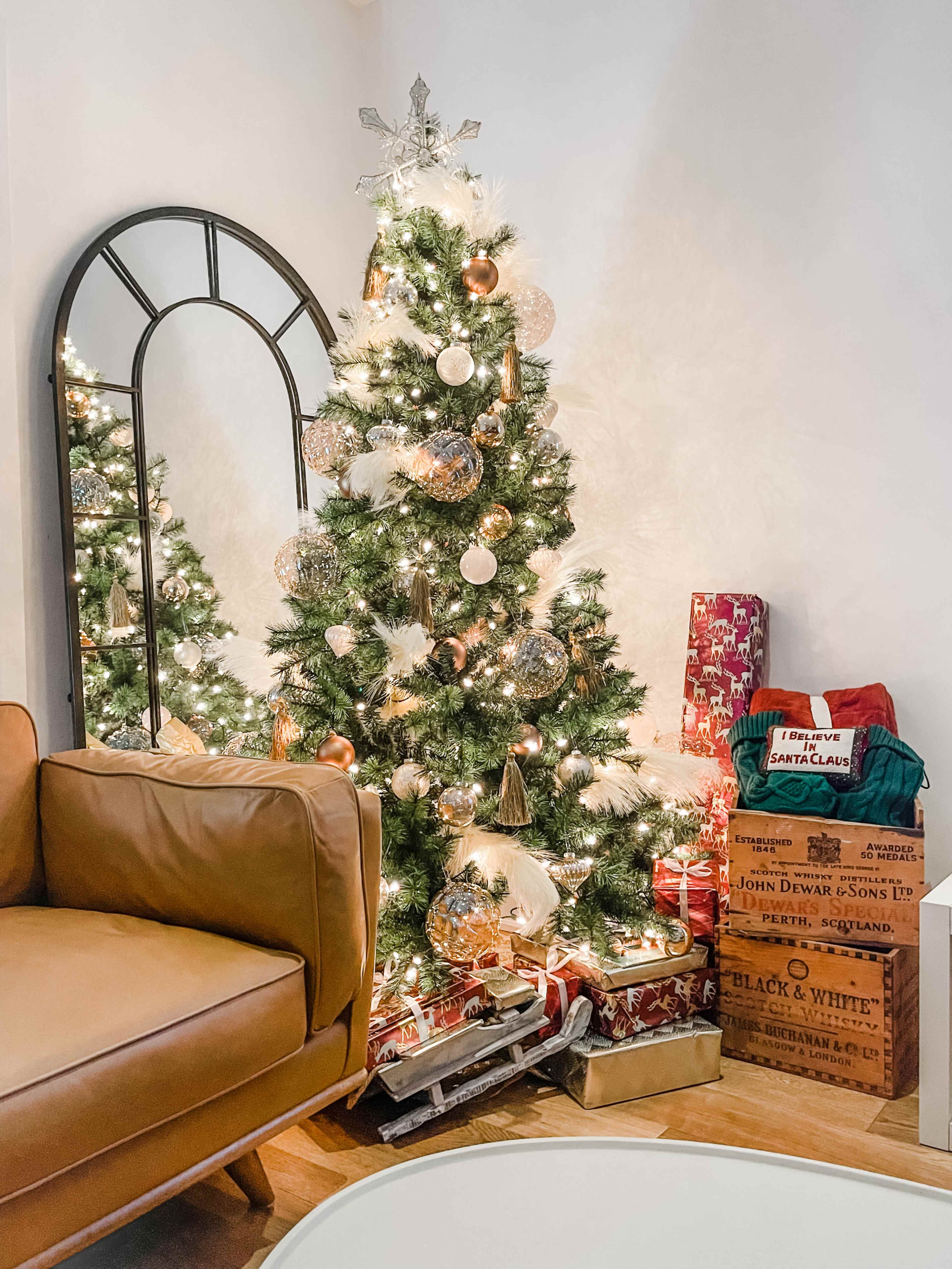A decorated Christmas tree stands in a cozy living room corner, surrounded by wrapped gifts and a large mirror.