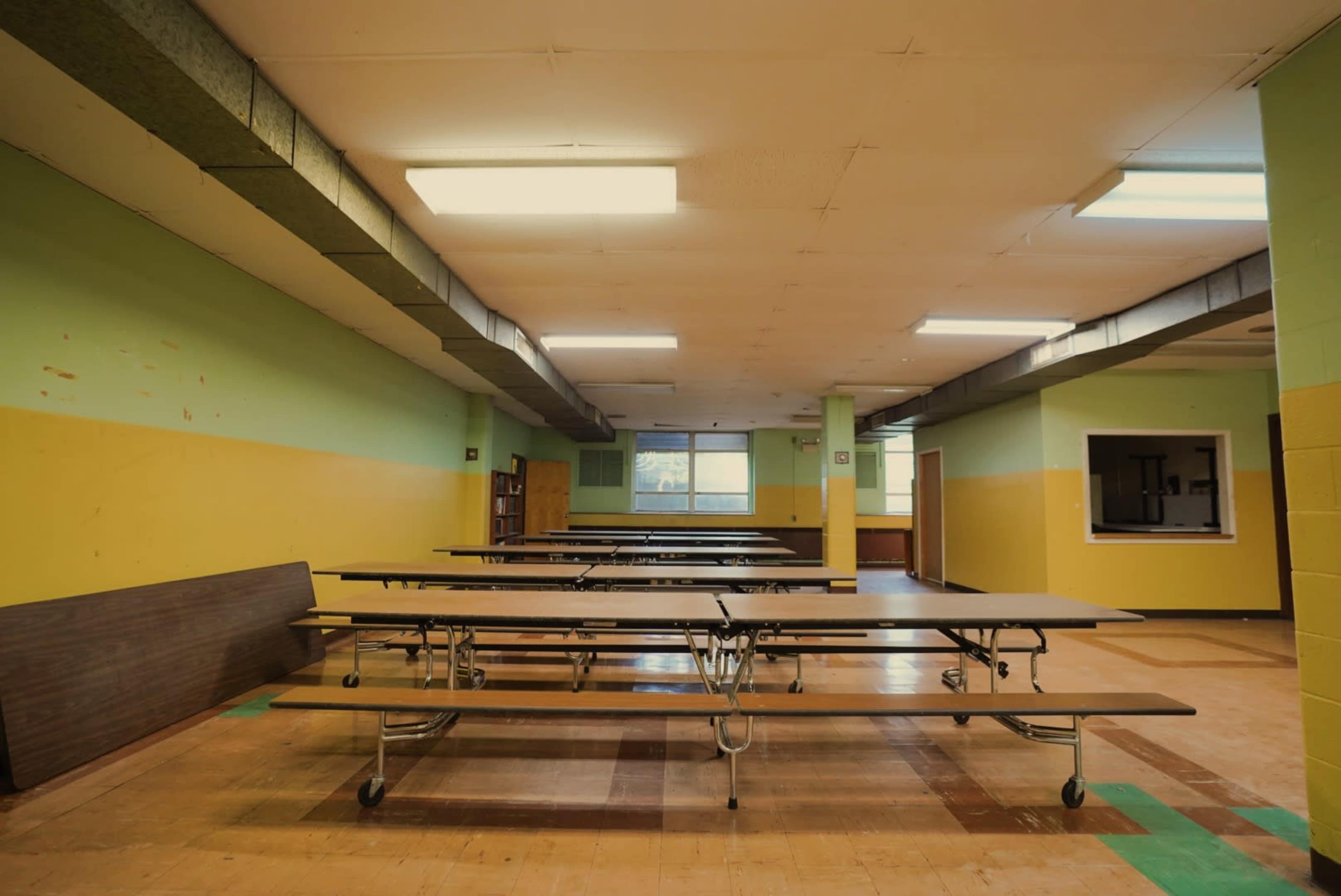 An empty school cafeteria with long tables arranged in rows and green and yellow walls.