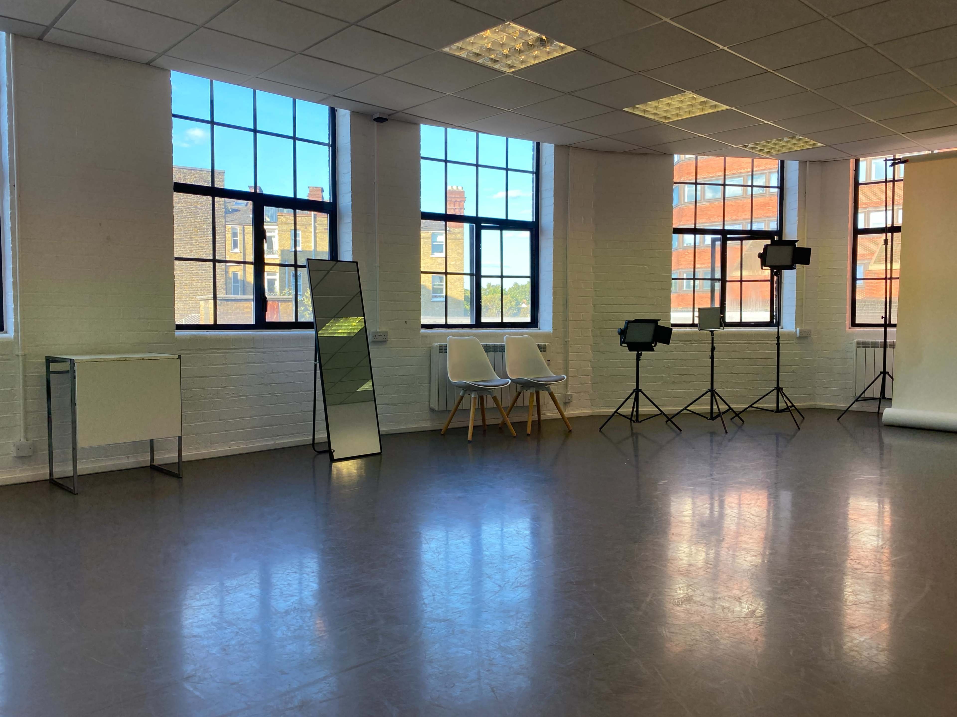 An empty studio space with large windows, two white chairs, a floor mirror, and lighting equipment on stands.