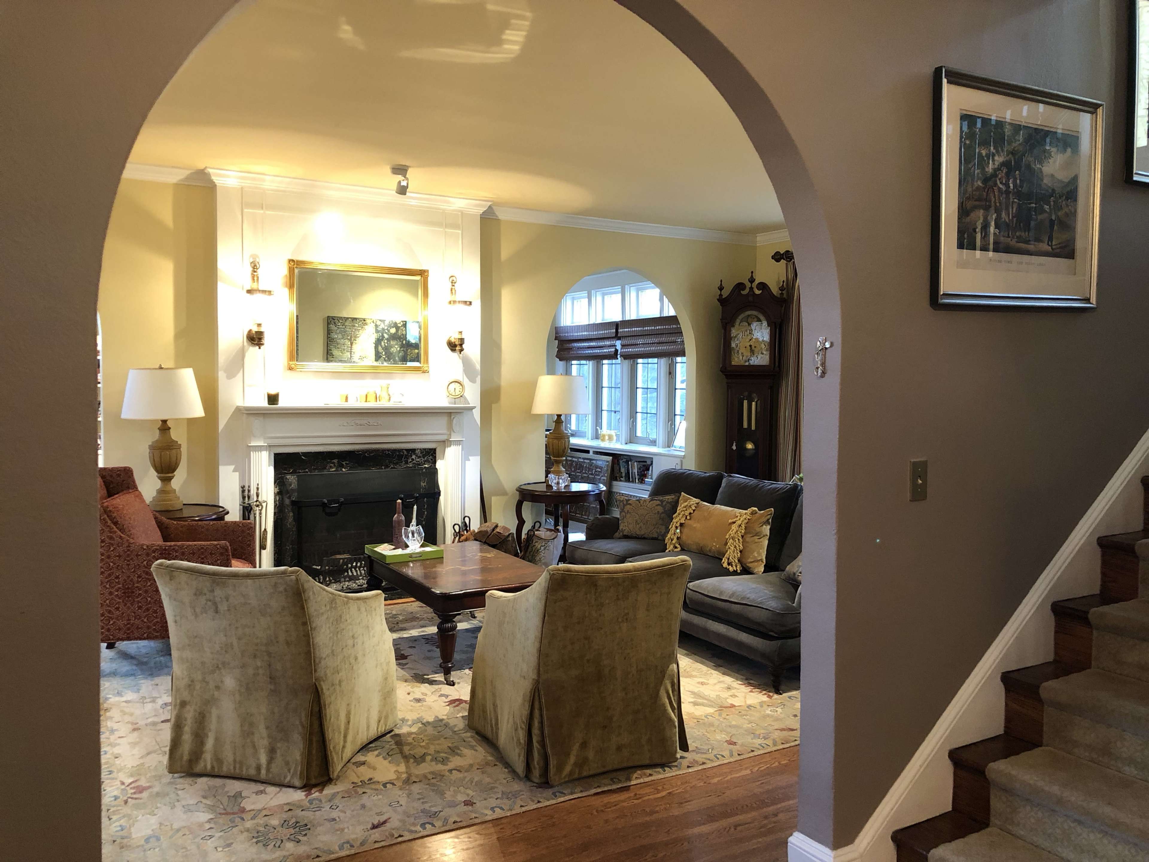 The image shows a living room with a fireplace, two upholstered chairs facing a coffee table, and a staircase visible in the background.