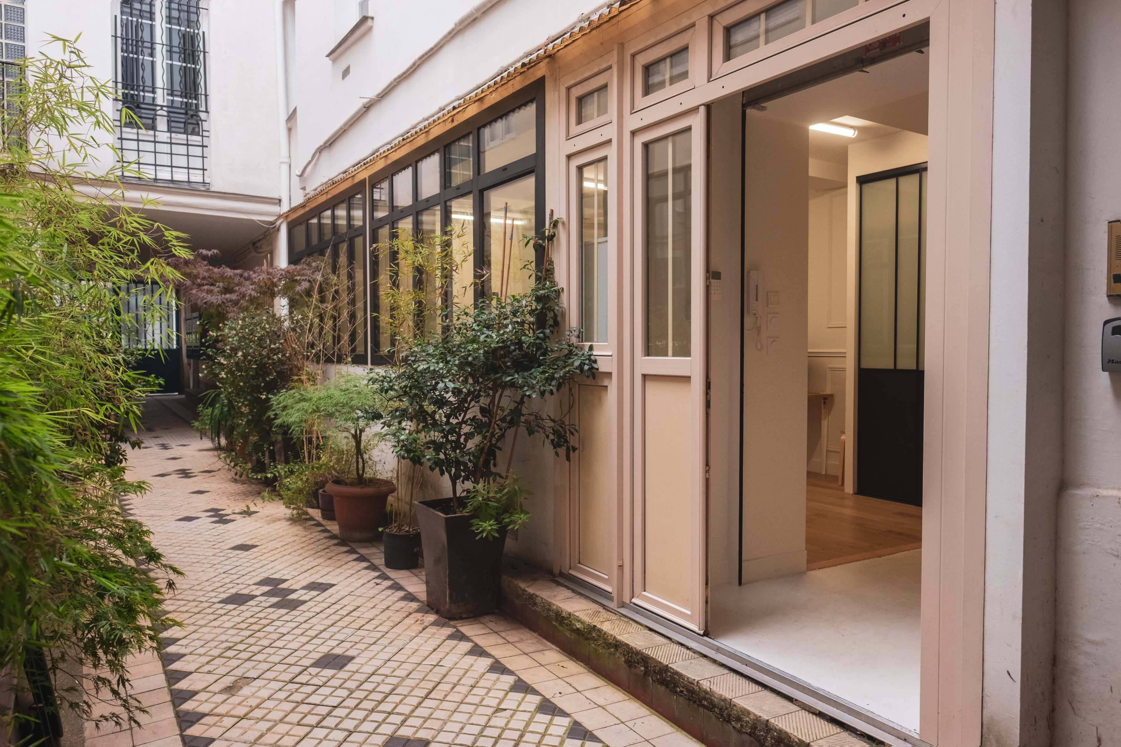 A narrow courtyard with a paved pathway lined with plants, leading to an entrance of a building featuring large glass windows and a light-colored door.