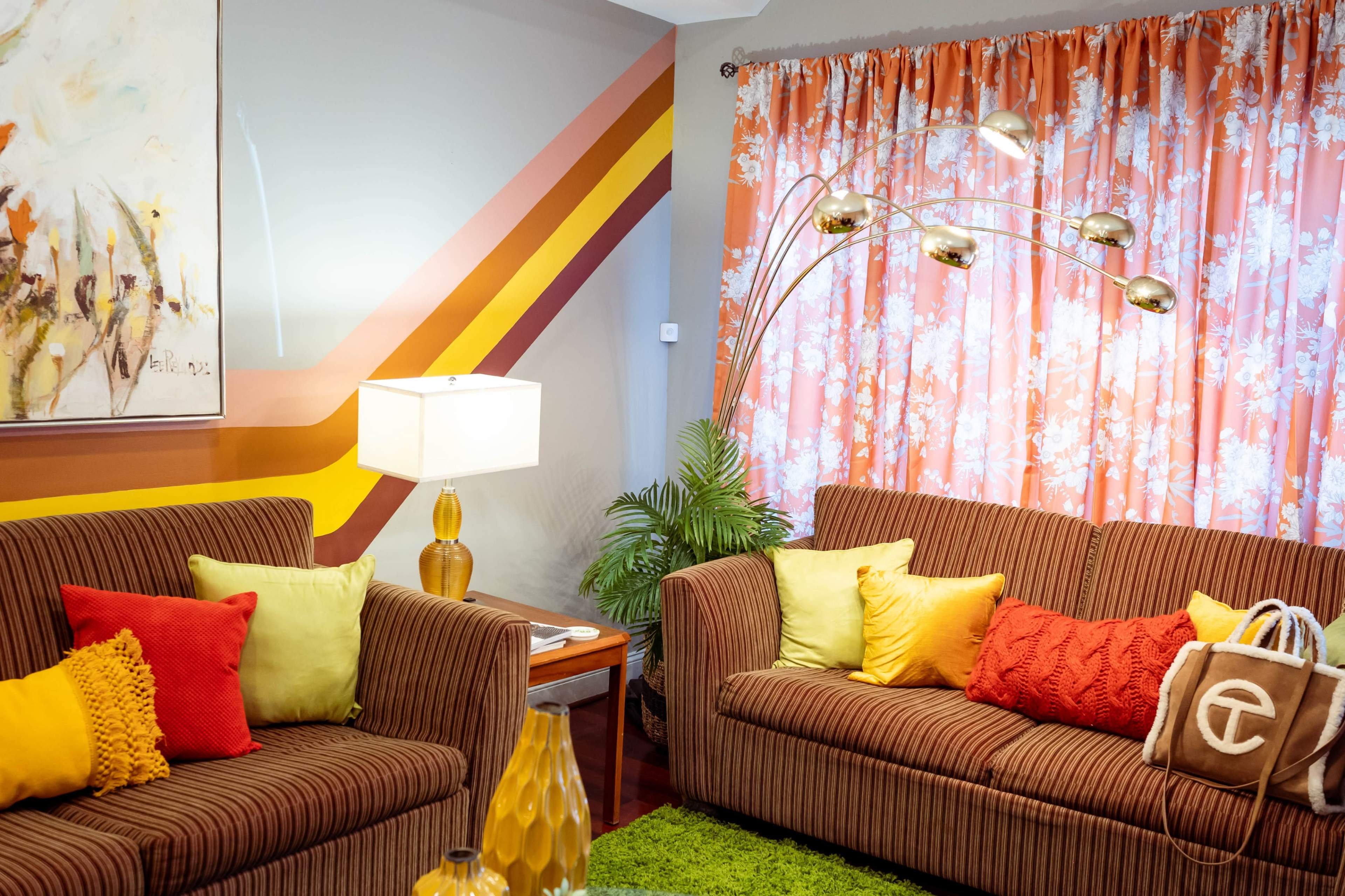 The living room features two striped sofas adorned with colorful cushions, a floor lamp, and vibrant floral curtains.